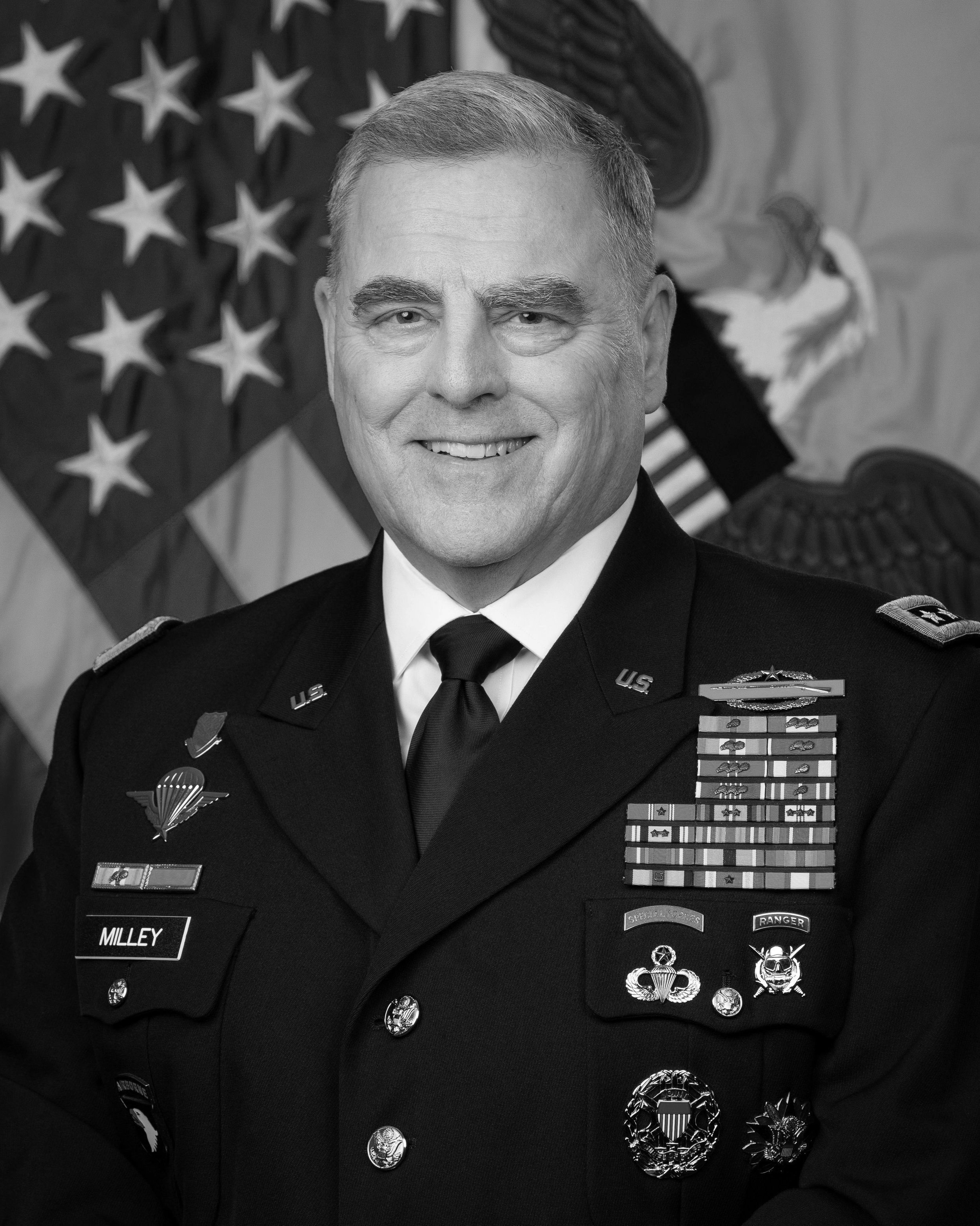 A smiling man in a United States military dress uniform, with numerous ribbons, medals, and badges, standing in front of an American flag and military insignia.