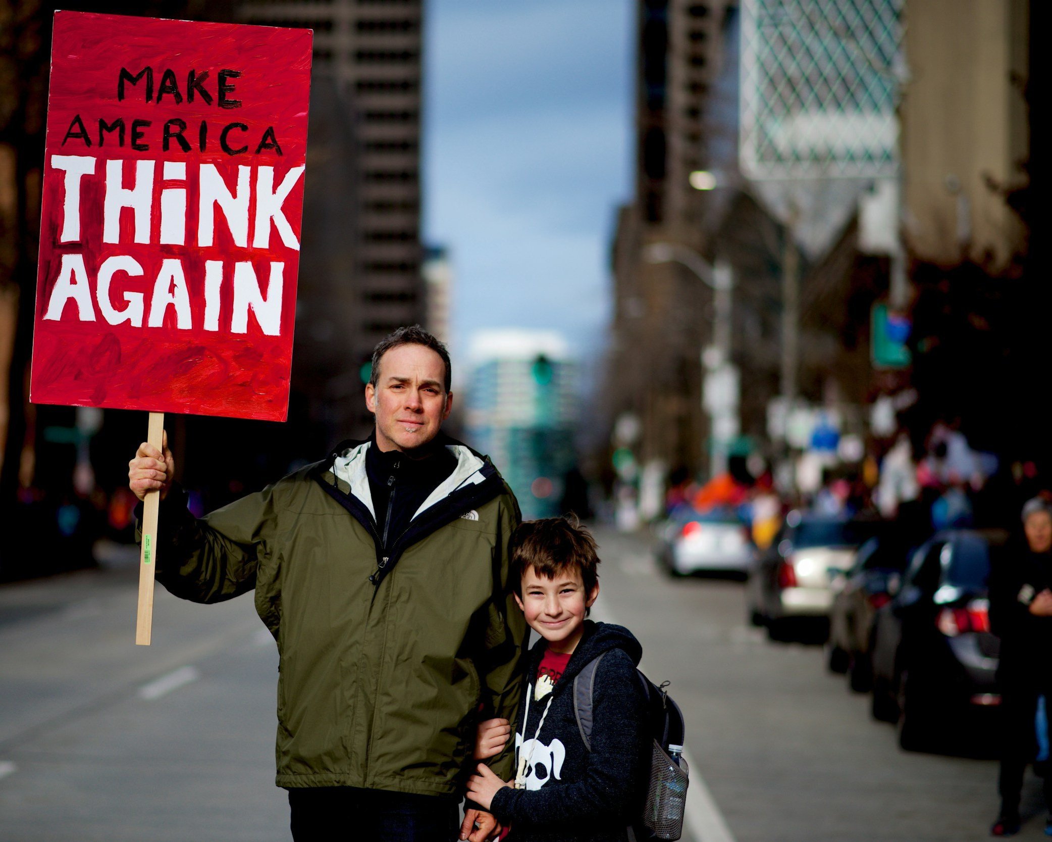 Man and boy standing on city street holding a red protest sign that reads, 'Make America Thank Again' with a cityscape in the background.