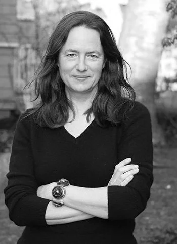 Black and white photo of a woman with shoulder-length wavy hair, wearing a black sweater and a wristwatch, standing outdoors with her arms crossed.