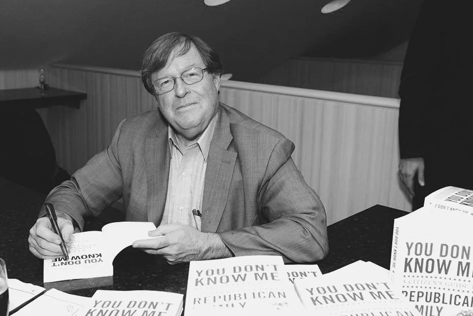 A man with glasses and light-colored hair, wearing a suit, is sitting at a table signing books. The table has multiple books titled "You Don't Know Me" and "Republican" on it.