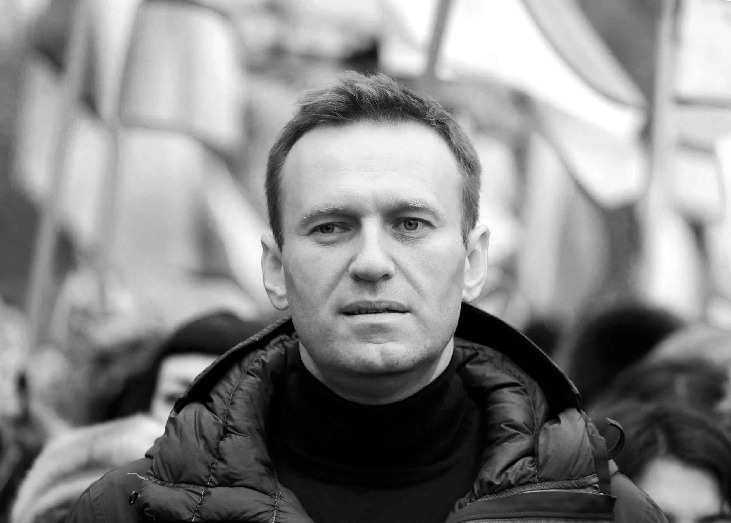 Black and white photo of a man with short hair wearing a puffy jacket, looking slightly to the side with a serious expression, outdoors with people in the background.