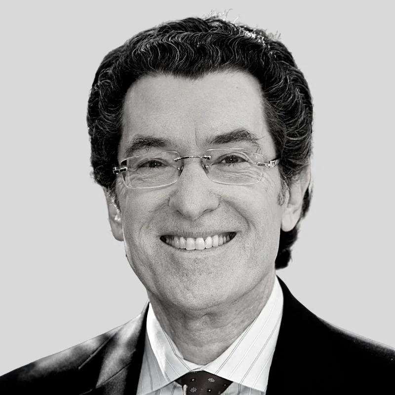 A black-and-white photo of a smiling man with dark, wavy hair, wearing glasses, a suit, dress shirt, and patterned tie.