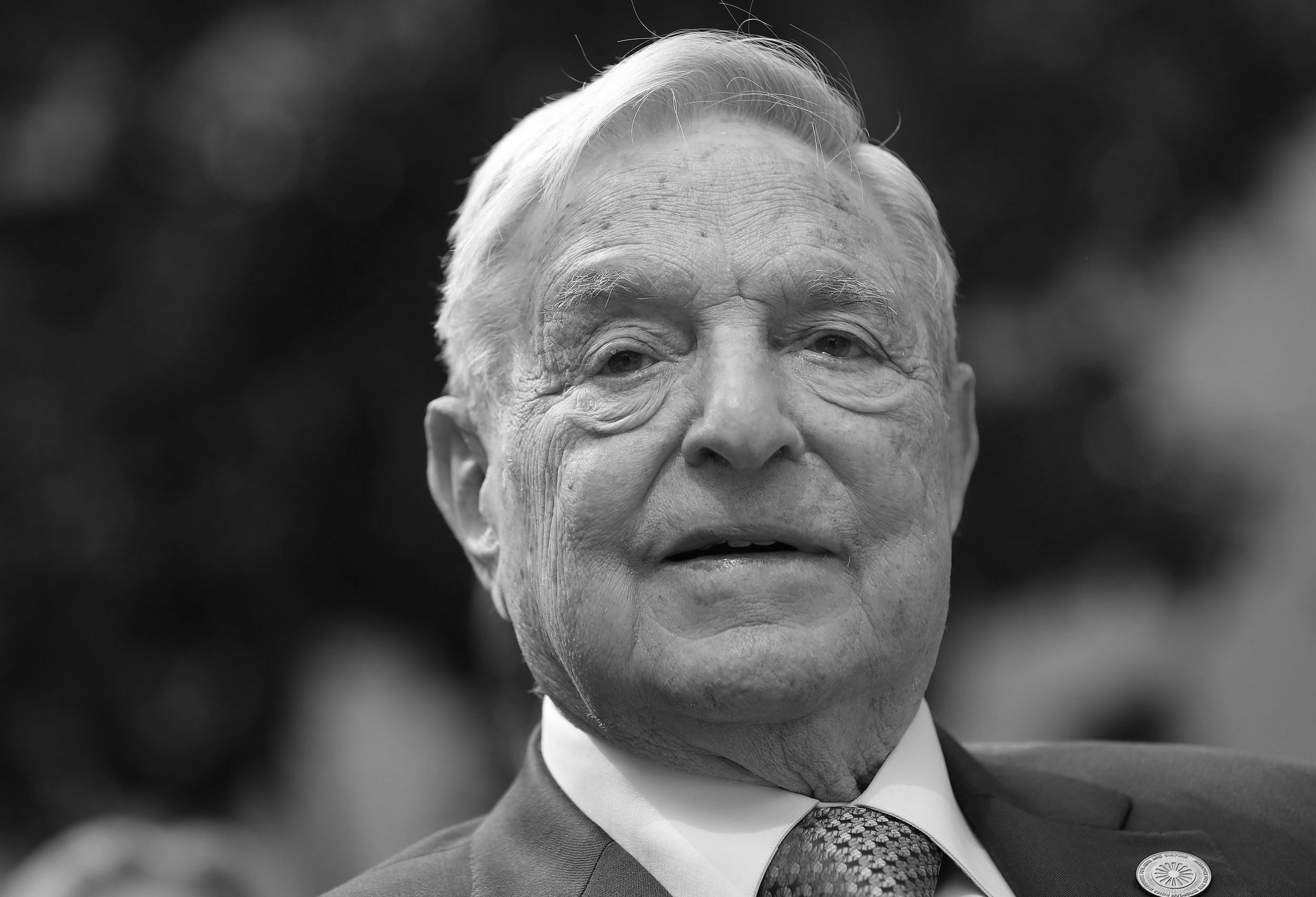 A close-up black-and-white photograph of an elderly man with white hair, wearing a suit and a patterned tie, looking directly at the camera with a serious expression.