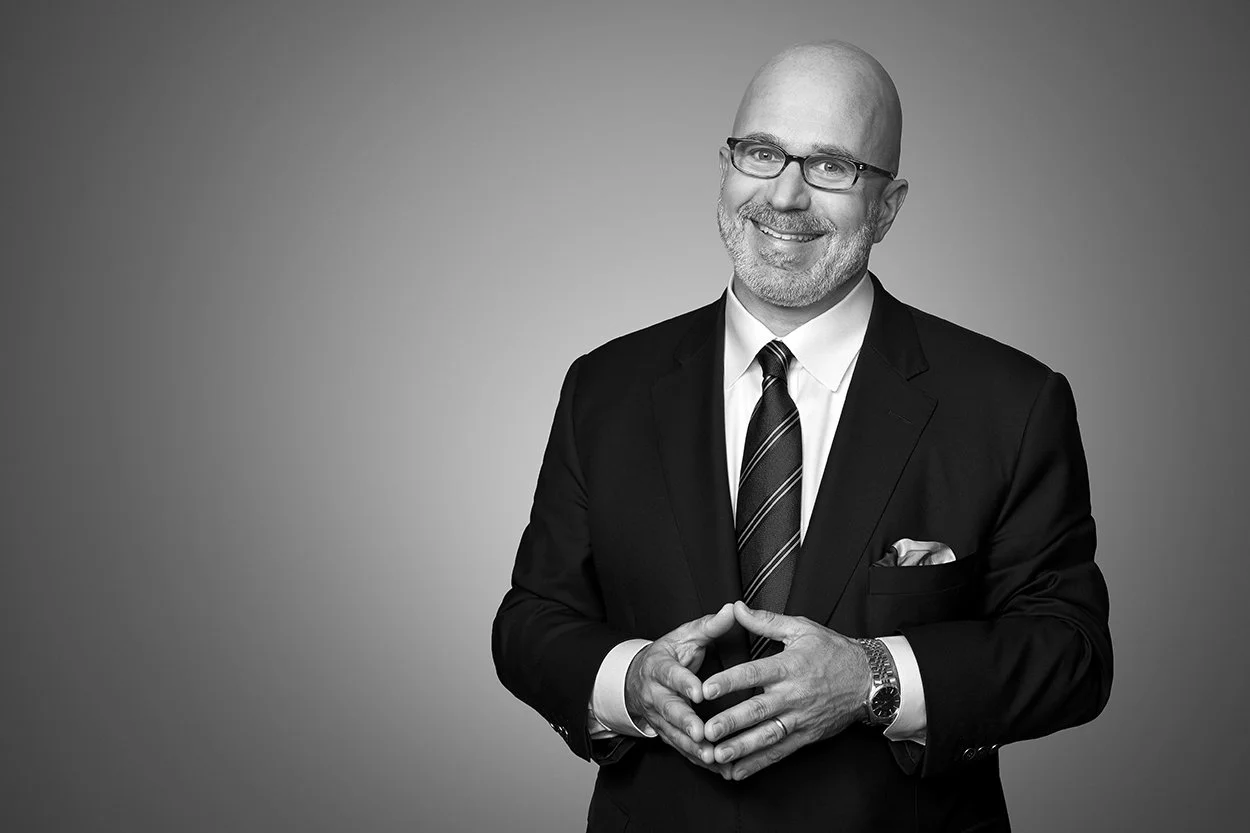A smiling, bald man with glasses, wearing a dark suit, white shirt, striped tie, and a watch, standing against a plain background.