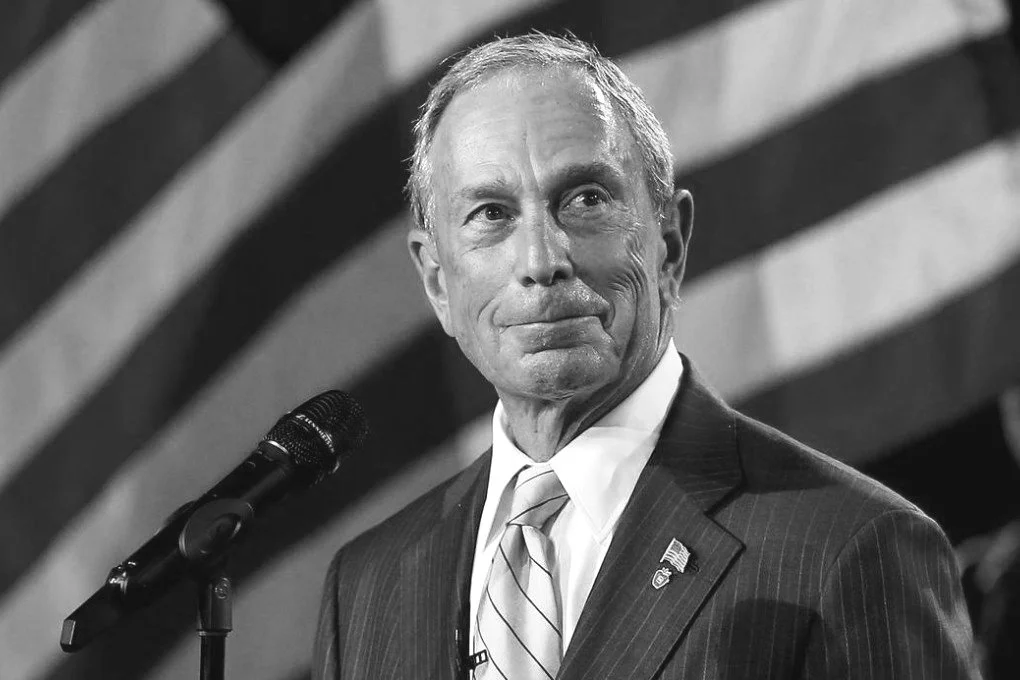 A black and white photo of a man in a suit and tie standing next to a microphone with an American flag in the background.