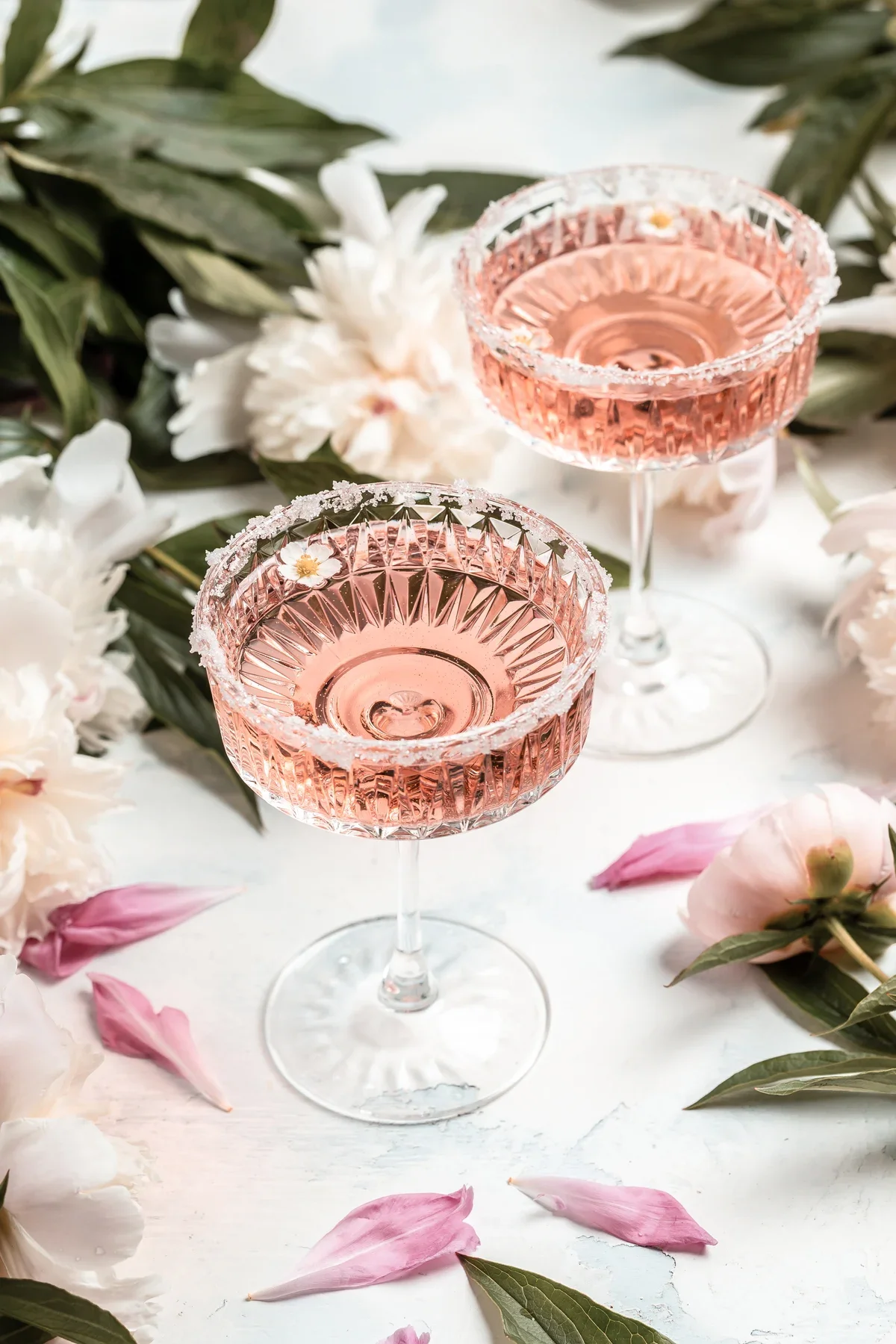 Two elegant pink cocktail glasses with sugar-rimmed edges, garnished with small white flowers, are surrounded by white and pink flowers and green leaves on a white surface.