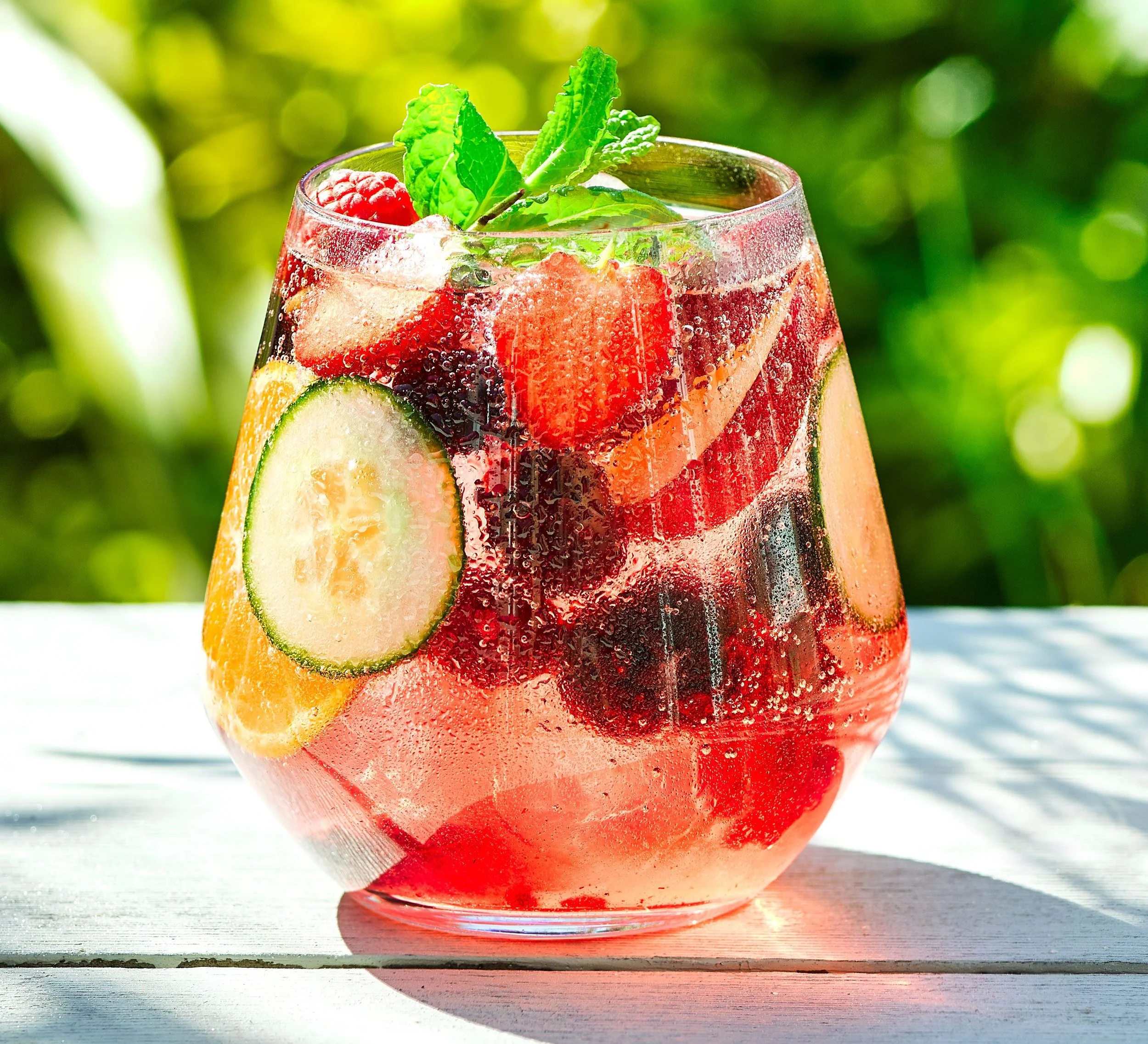 A glass of fruit-infused soda with ice, slices of cucumber and orange, and fresh raspberries, garnished with mint leaves.