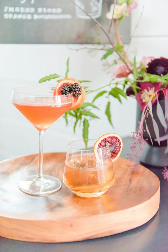 Two pinkish-orange cocktails with fruit garnishes on a wooden tray, with a purple flower vase and green plants in the background.