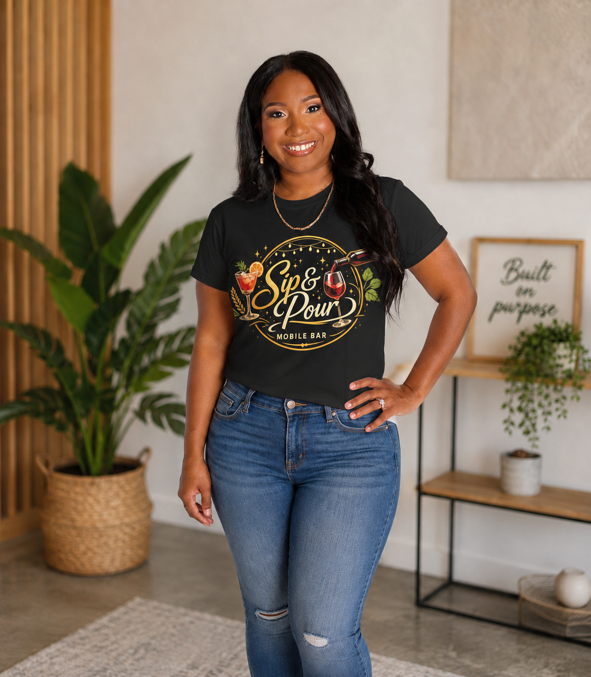 Woman standing indoors in front of a wooden and beige wall, wearing a black t-shirt with a logo and jeans, smiling at the camera.
