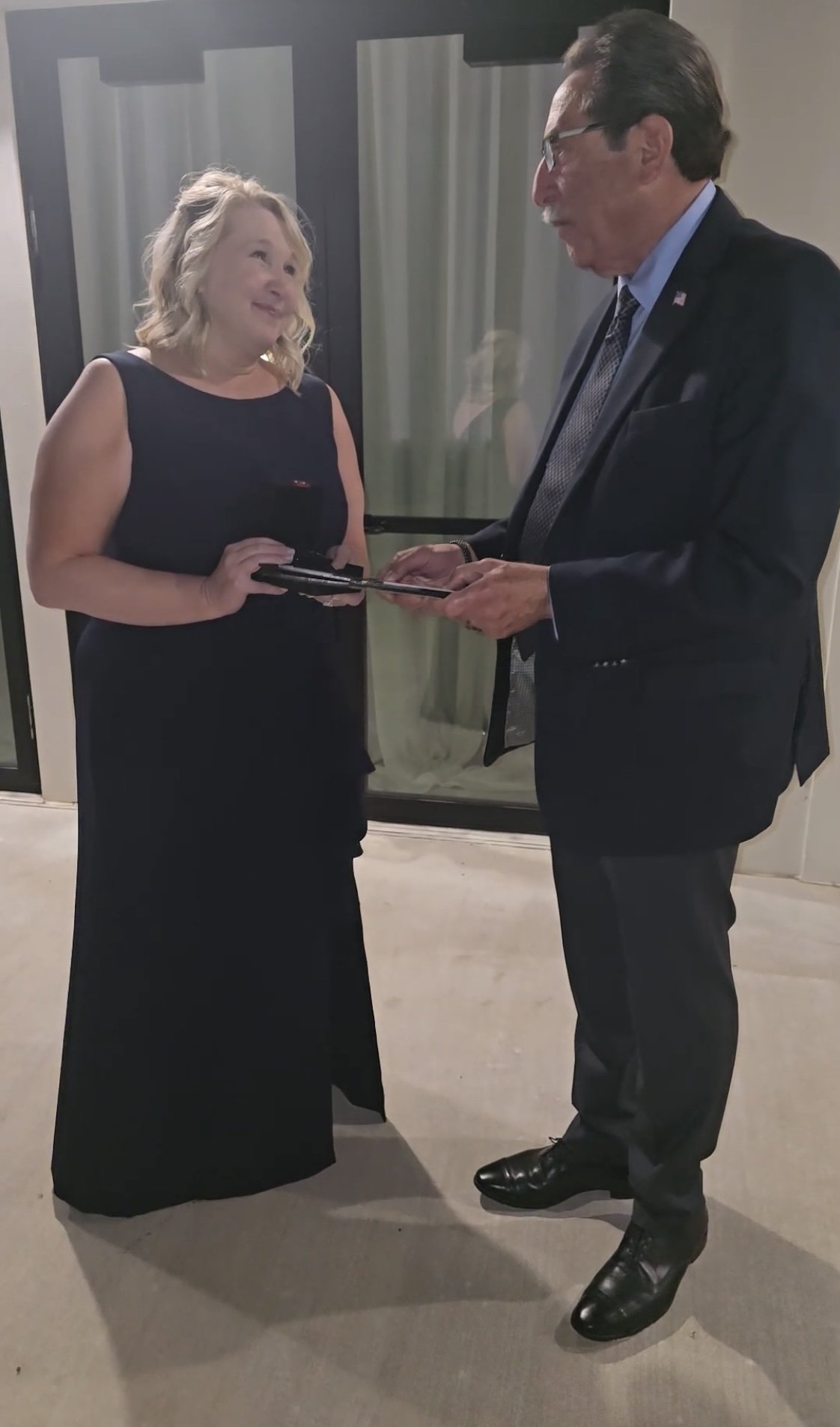 A woman in an elegant black dress holding a small box and a man in a suit exchanging items outdoors.