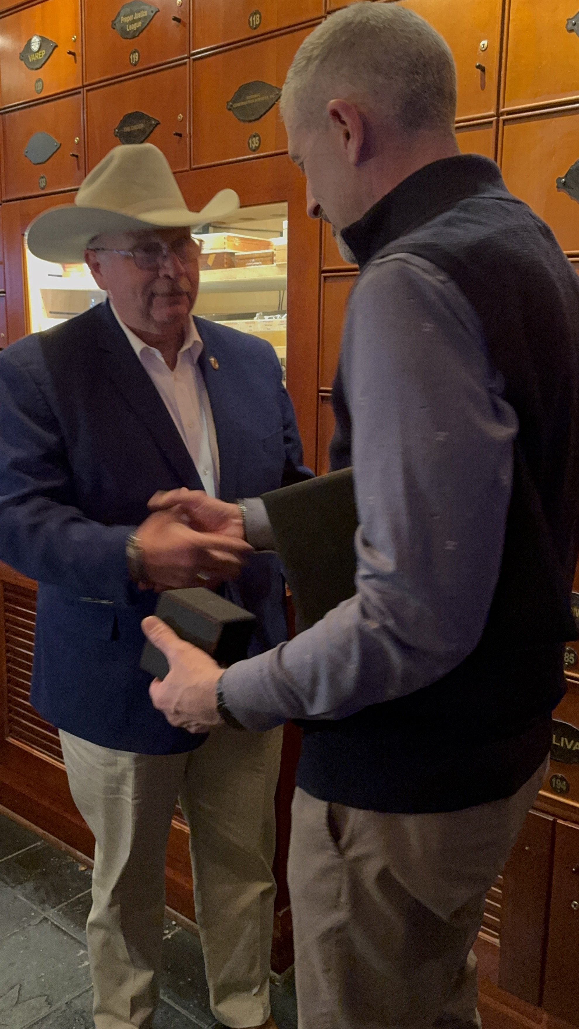 Two men shaking hands in a formal setting, one wearing a cowboy hat and the other holding a box.
