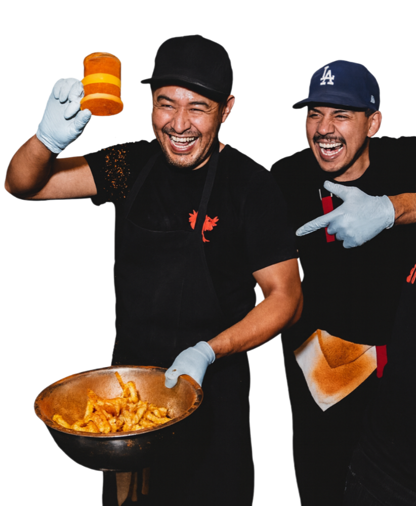 Two smiling men wearing black T-shirts, aprons, and baseball caps, one holding a frying pan with fried food and the other holding a container of dipping sauce, both wearing gloves and posing enthusiastically.