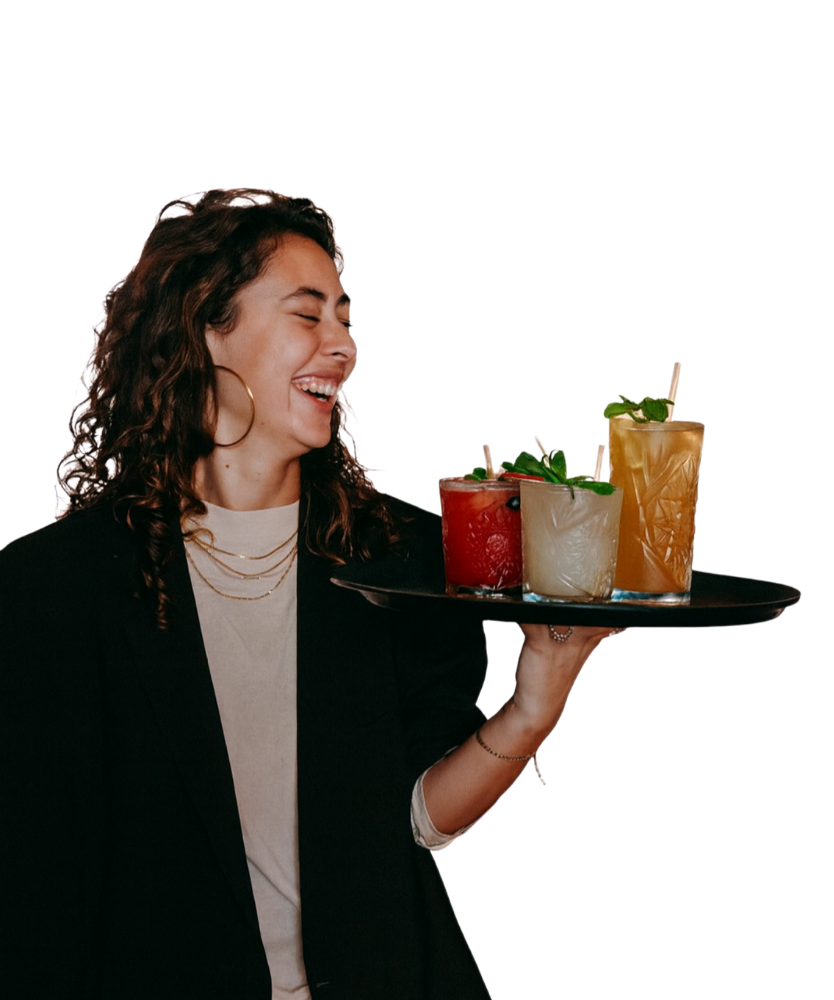 A woman with curly hair, wearing a black blazer and white shirt, smiling and holding a tray with three colorful cocktails garnished with mint leaves.