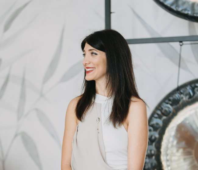 Smiling woman with dark hair in a white sleeveless top, posing indoors with a blurred floral patterned wall and studio lighting equipment in the background.