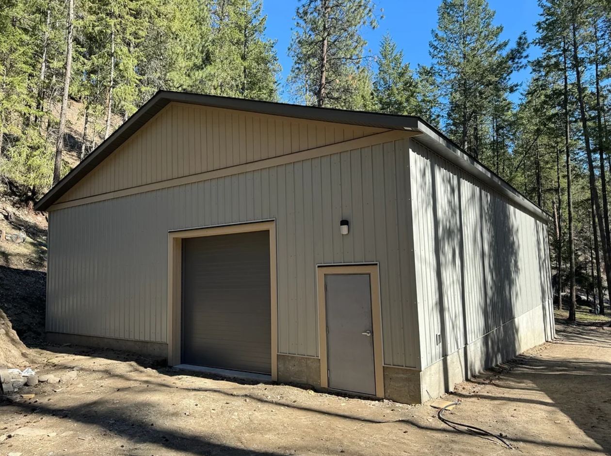 A beige metal storage building with a large roll-up door and a smaller pedestrian door, situated in a wooded area with tall trees and dirt ground.
