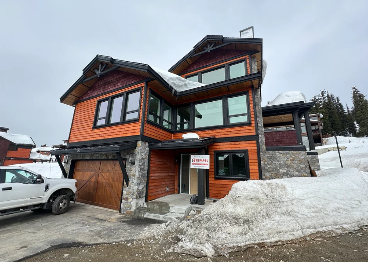 Modern multi-story house with wooden and stone exterior, large windows, snow on the ground, and a white pickup truck parked outside.