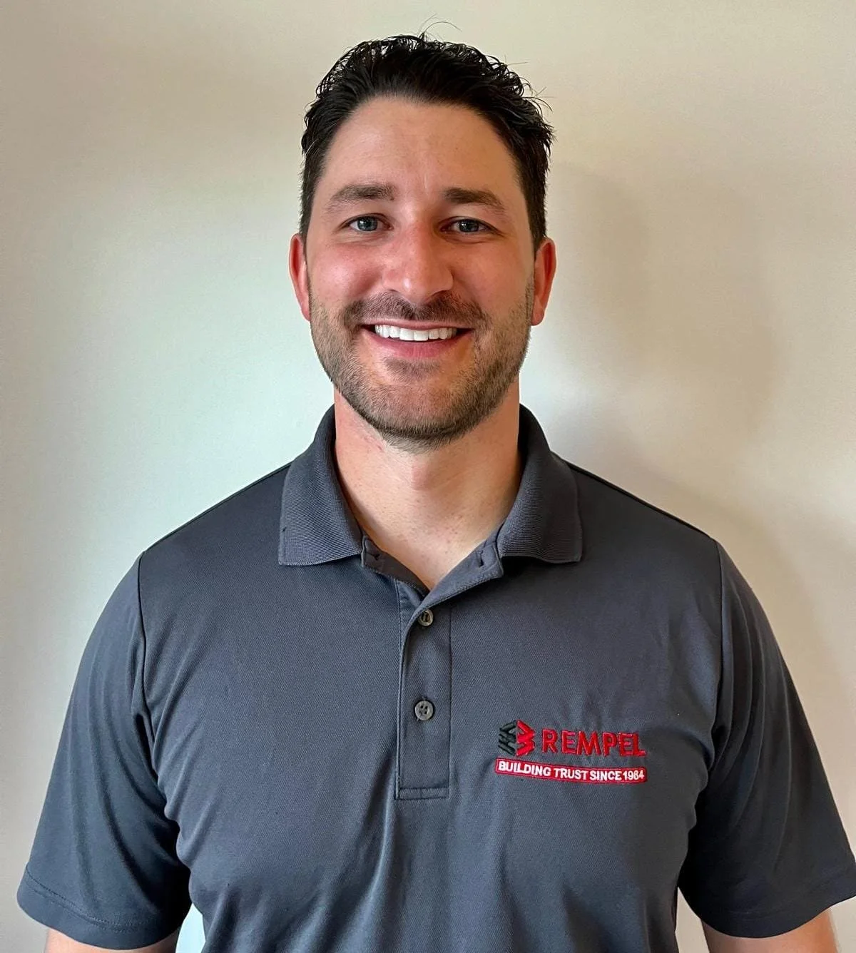 A man smiling, wearing a dark gray polo shirt with a REMPEL logo on the left chest and a light-colored background.