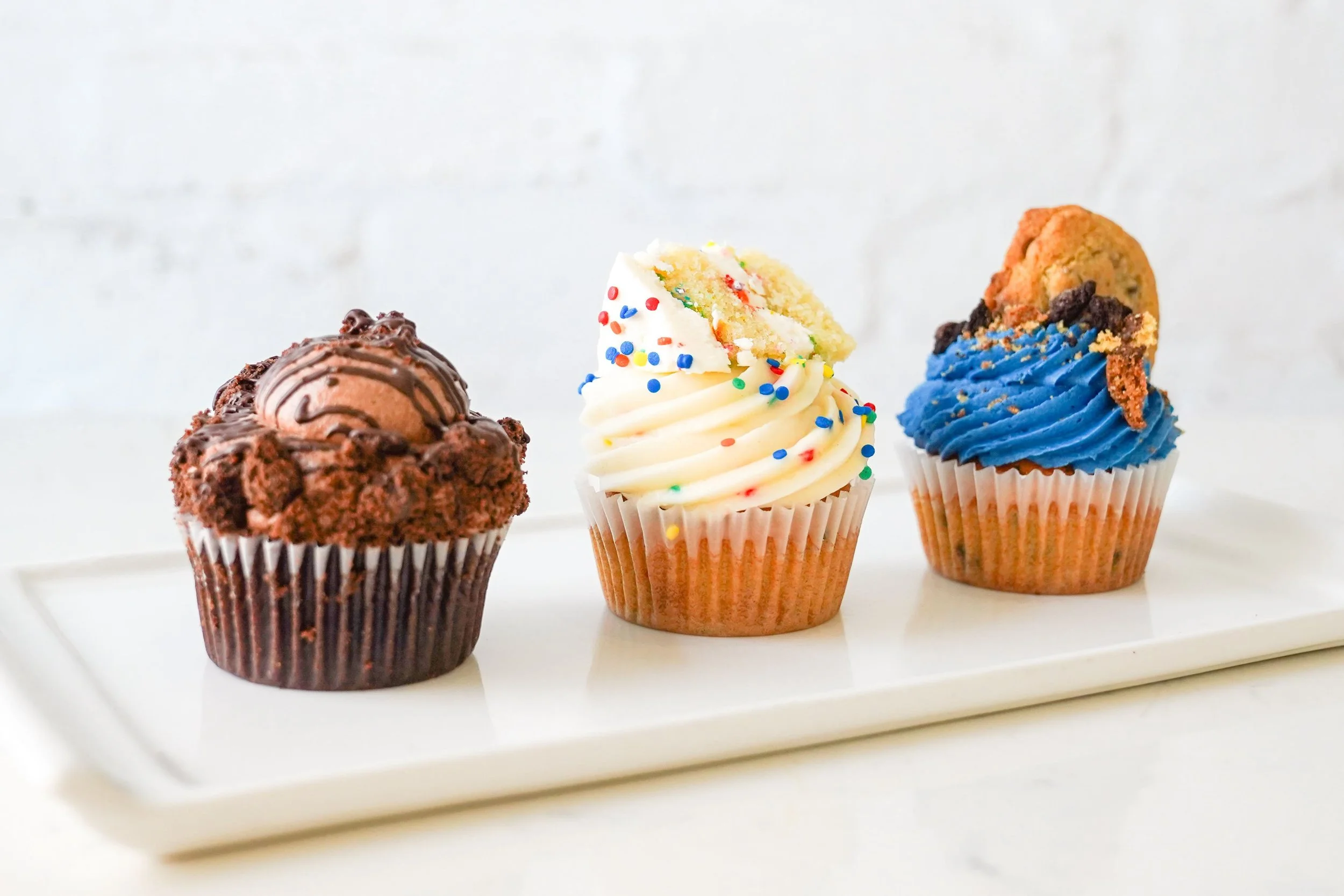 Variety of cupcakes on a white tray, featuring chocolate, vanilla with sprinkles, and blue frosted cookies and cream flavors.