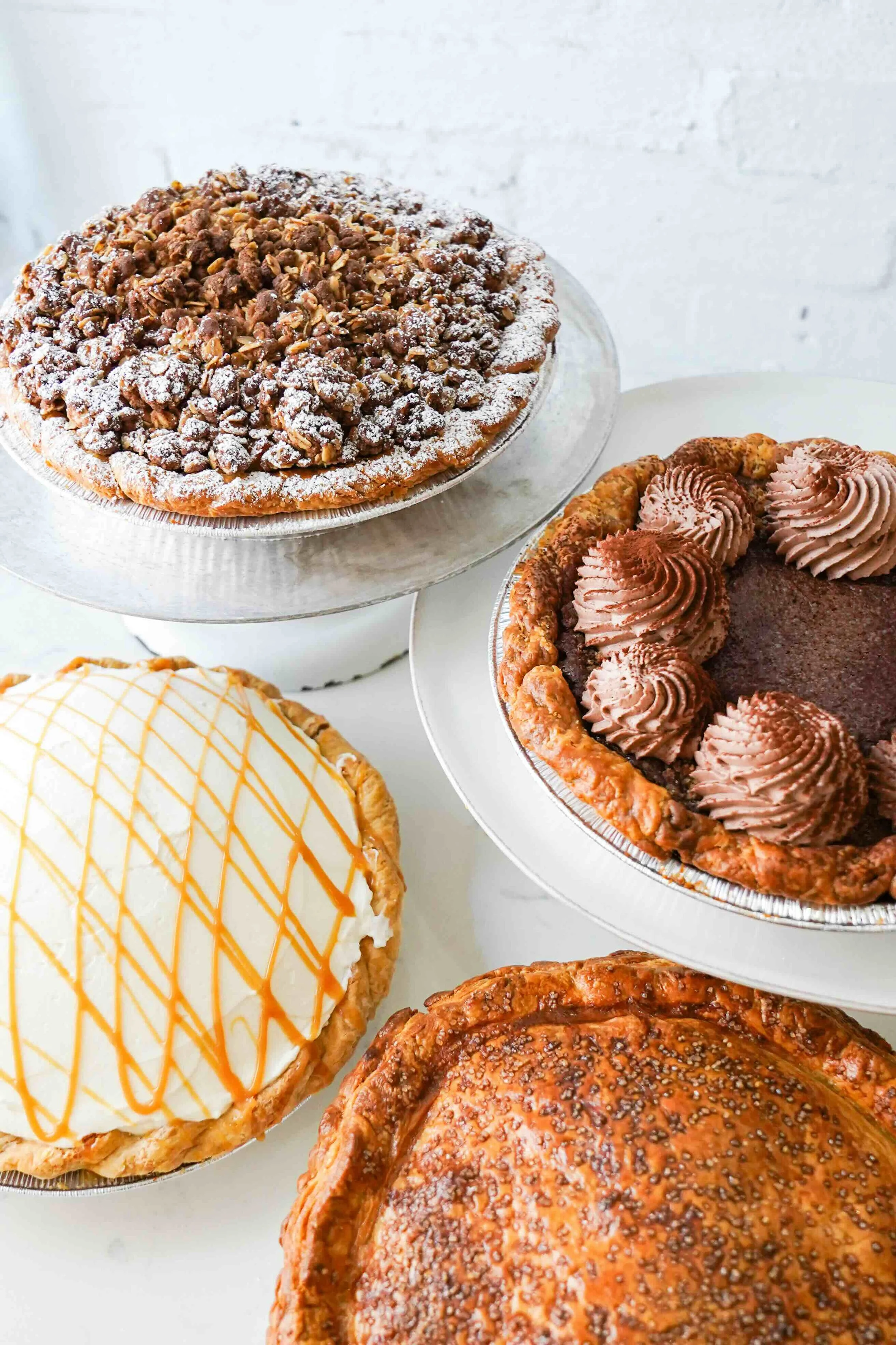 Assorted pies on stands, including crumb, whipped cream, and chocolate decorated pies.