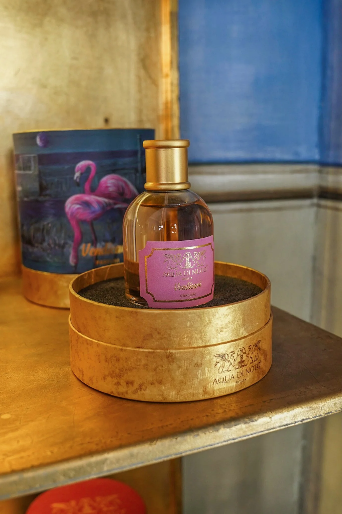 Elegant perfume bottle with gold cap and pink label on a gold base, next to a decorative box featuring a flamingo.