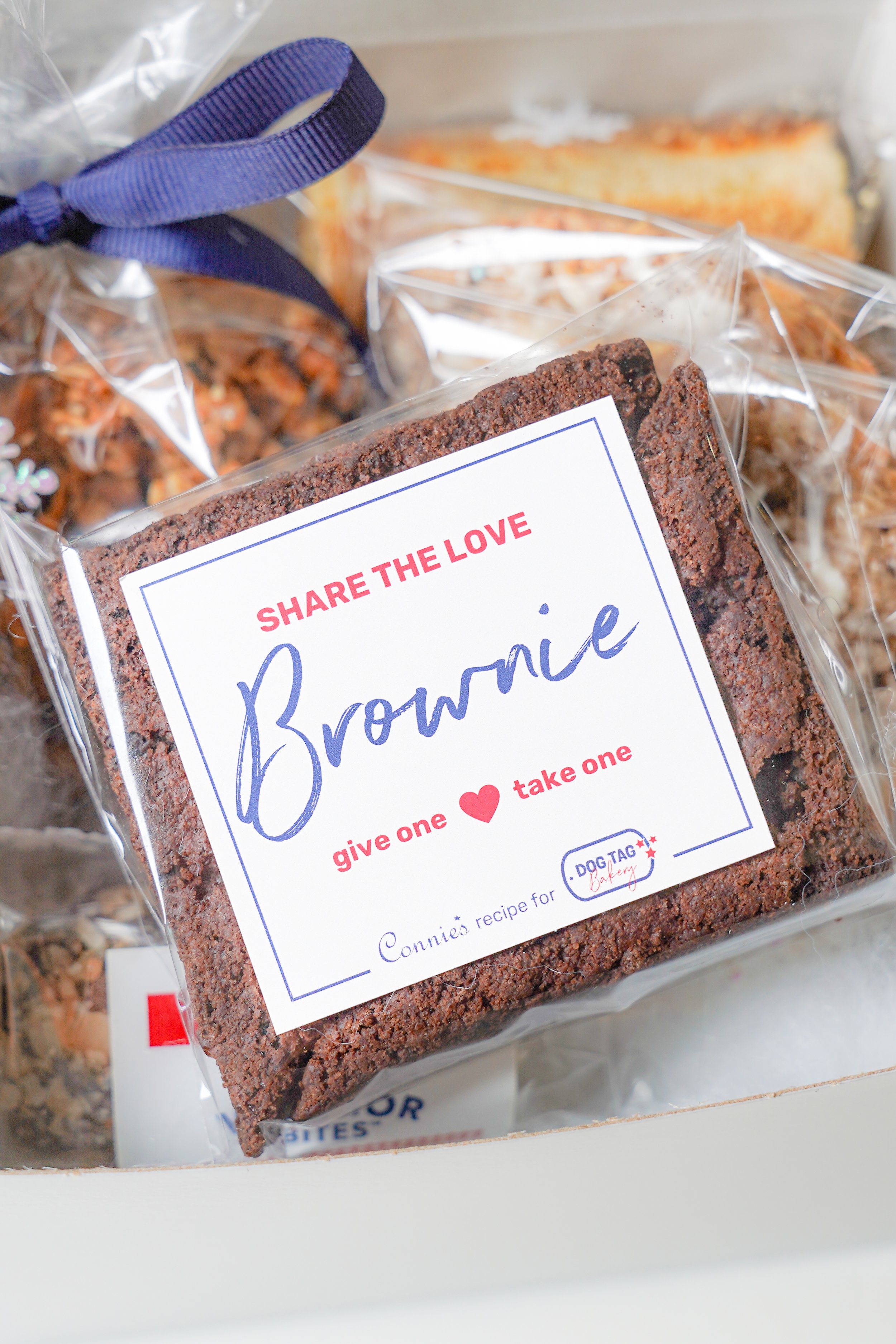 Close-up of a packaged brownie with a label reading 'Share the Love Brownie, give one ♥️ take one'. A blue ribbon is nearby.