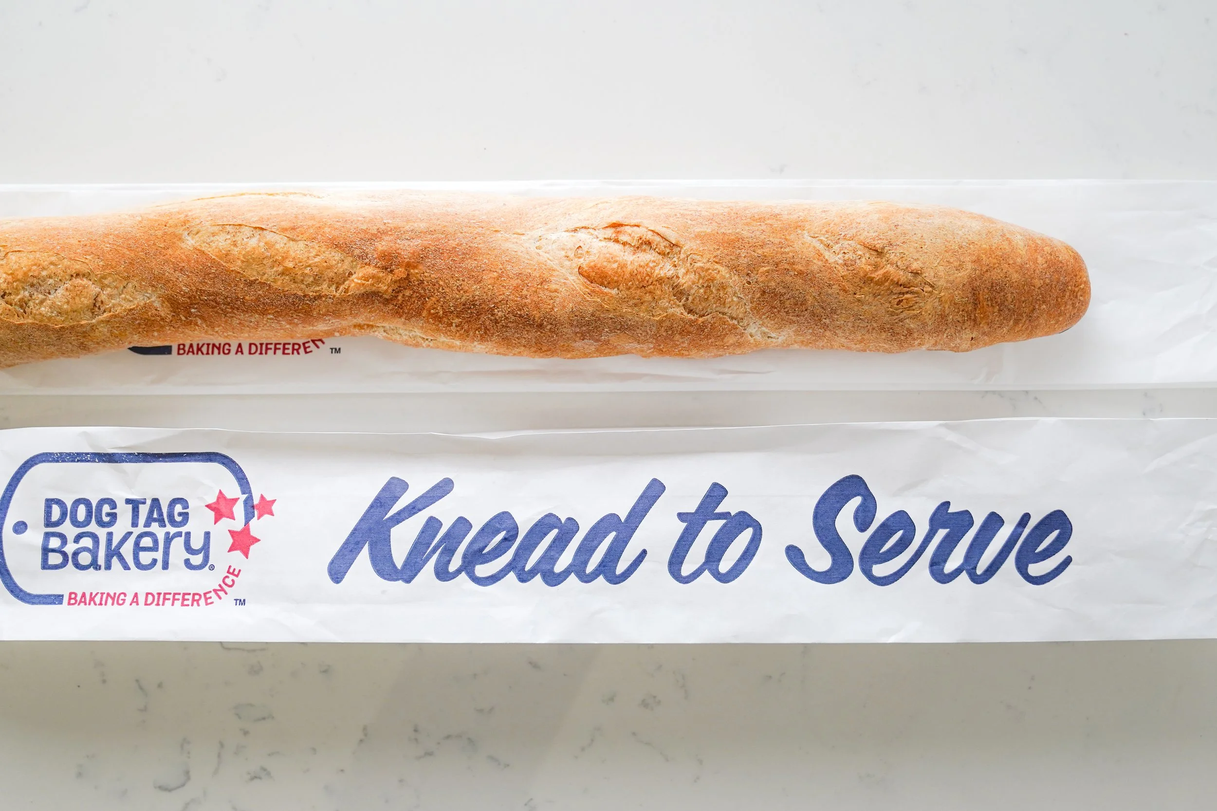A baguette from Dog Tag Bakery on a wrapper with "Knead to Serve" written on it.