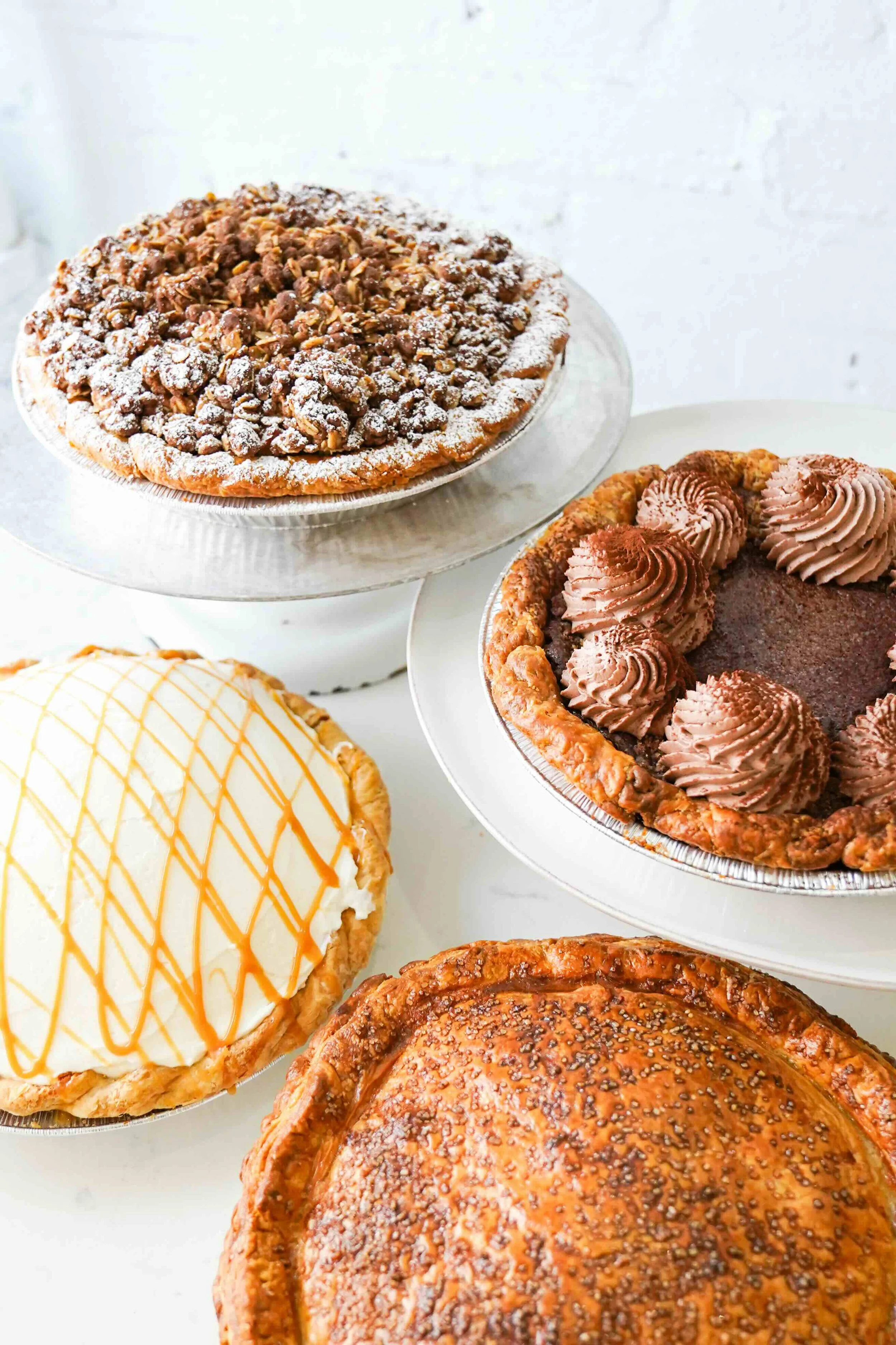 Assorted pies on cake stands, including a crumble top pie, a chocolate cream pie, a caramel drizzled pie, and a traditional pie with a speckled crust.