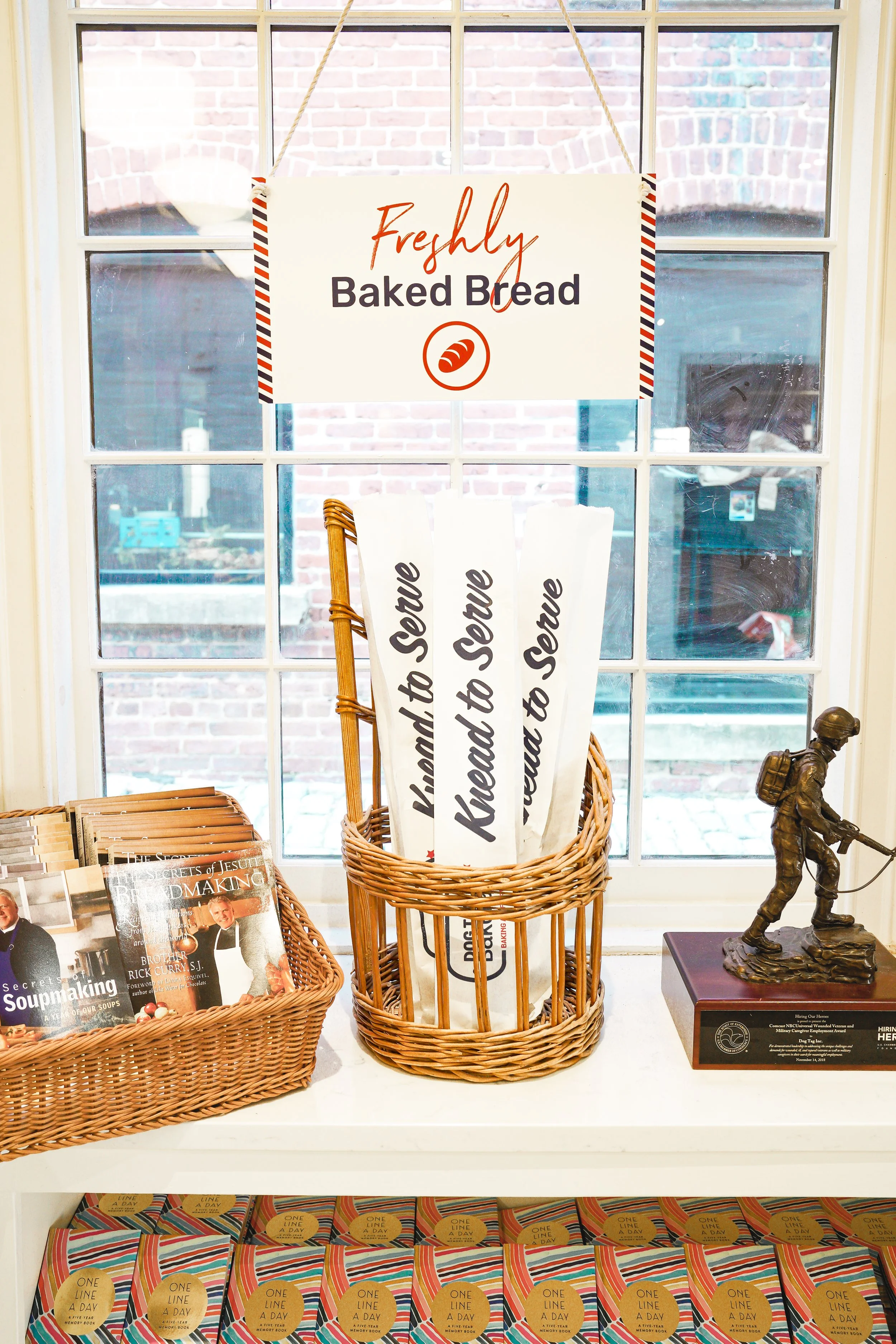Display of freshly baked bread sign, wicker baskets with bread sticks labeled 'Knead to Serve,' cookbooks, and small statue.