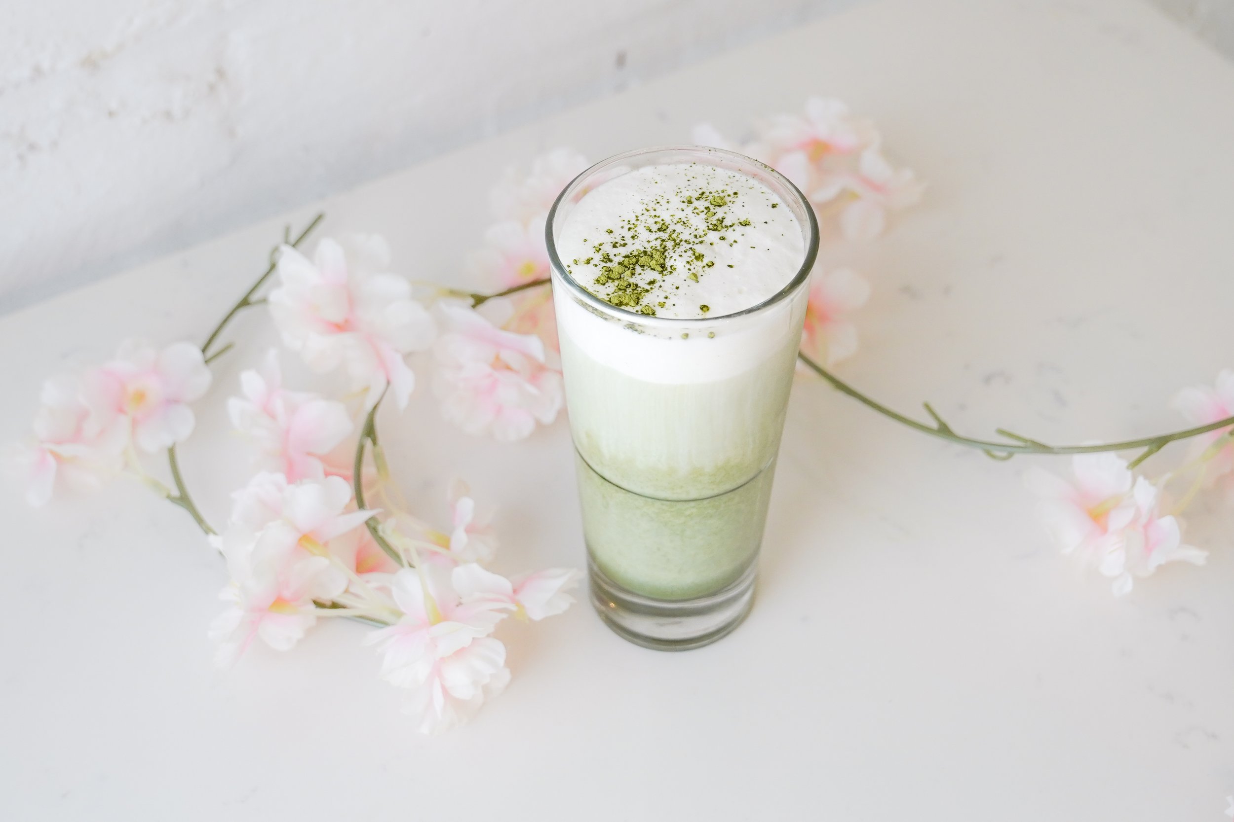 Matcha latte in a glass with foam and green powder on top, surrounded by pink and white flowers on a white table.