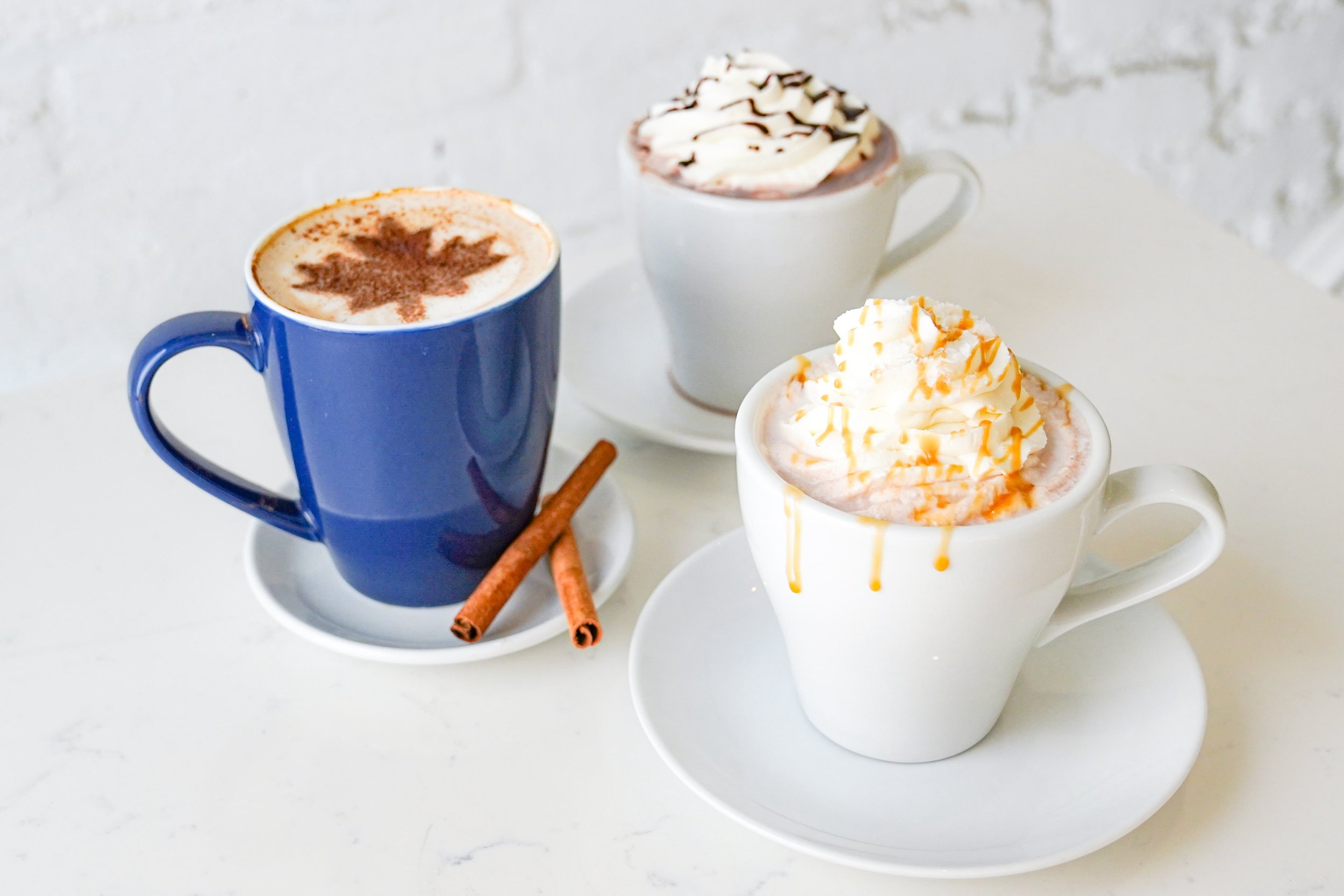 Three mugs of hot beverages; blue mug with leaf pattern, white mug with whipped cream and chocolate drizzle, and another white mug with whipped cream and caramel drizzle, on a white background.