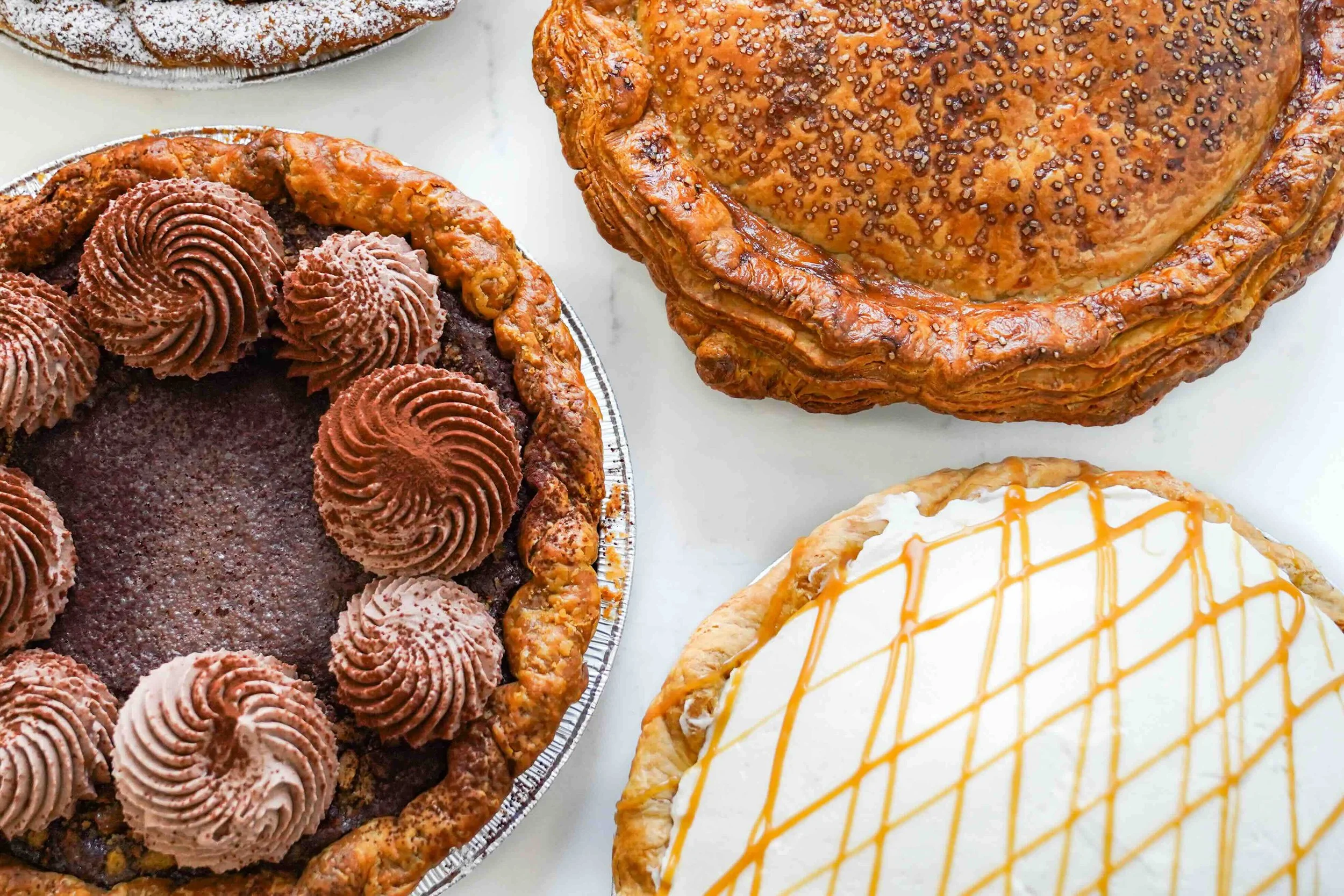 Assorted pies including chocolate mousse, caramel drizzle, and traditional topped pies on a white surface.