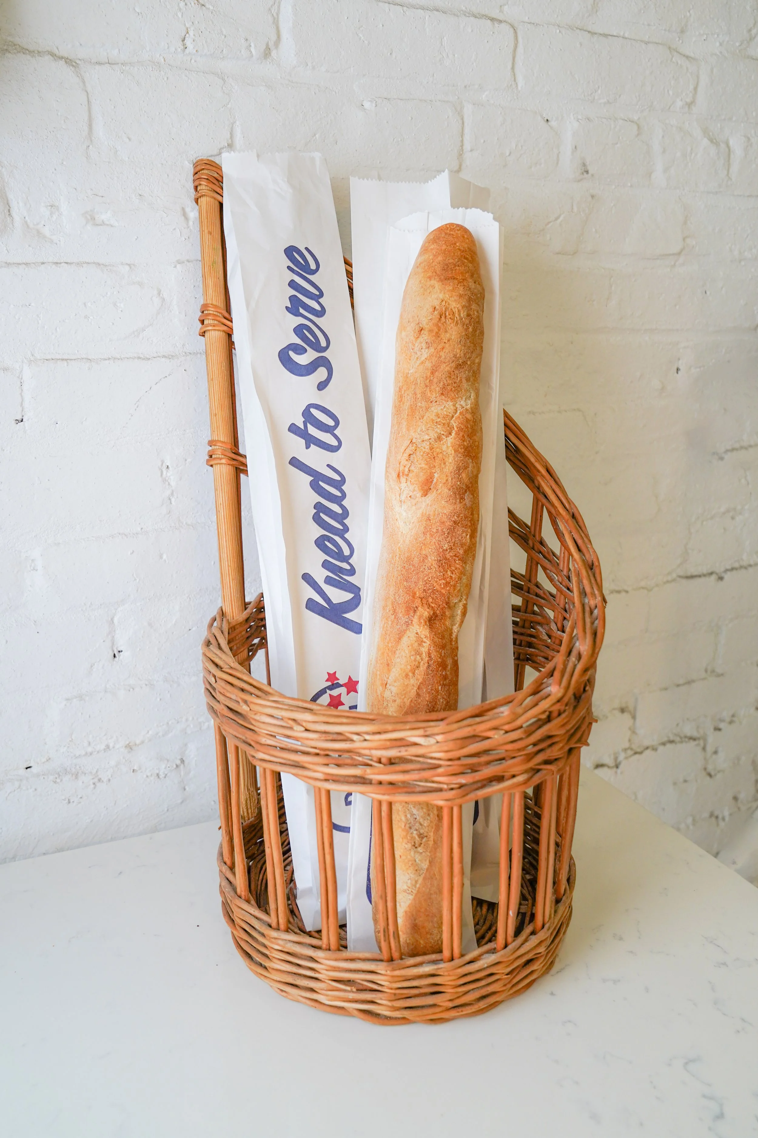 Baguette in a basket with 'Knead to Serve' paper label, white brick wall background.
