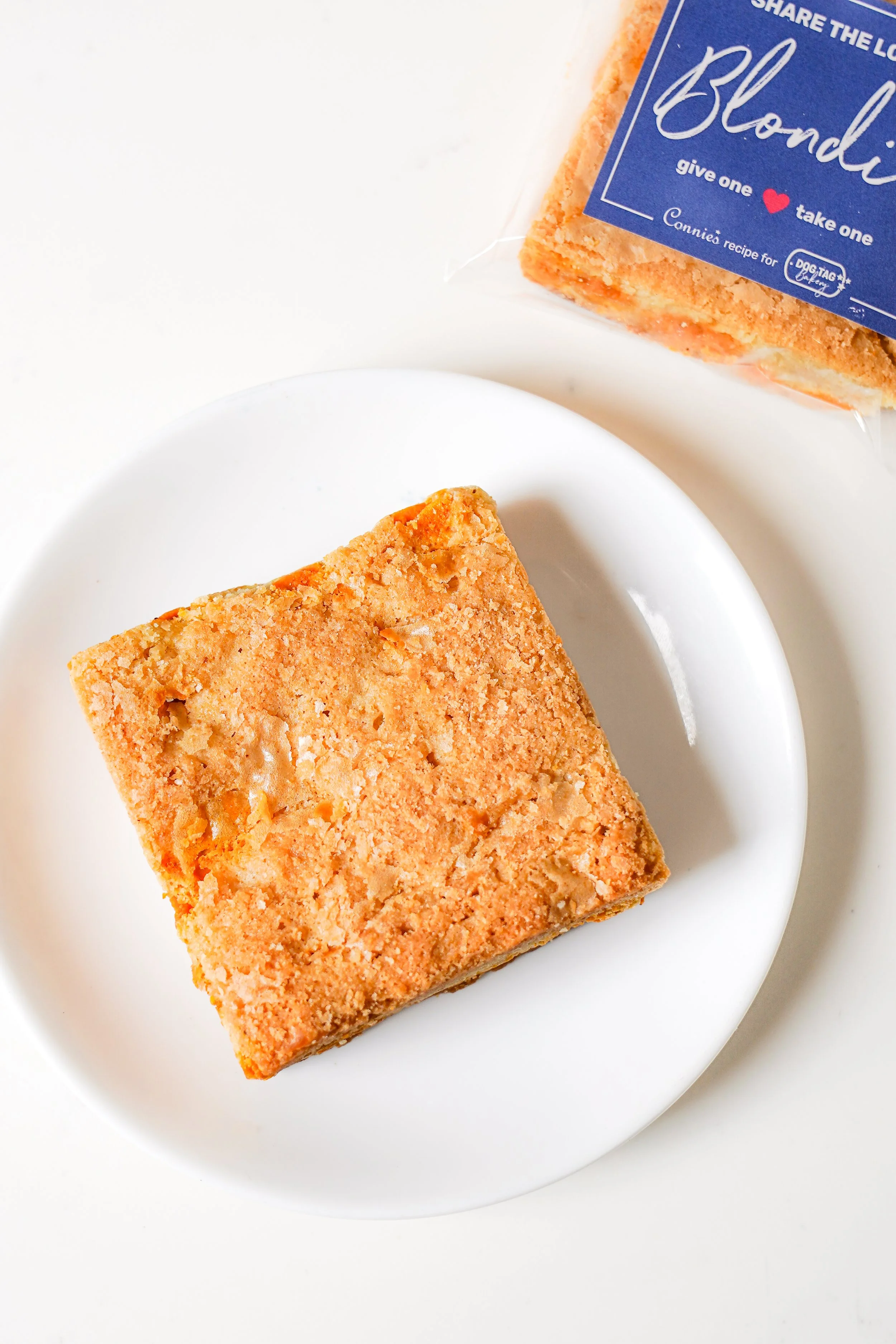 Square blondie dessert on a white plate with packaged blondie beside it.