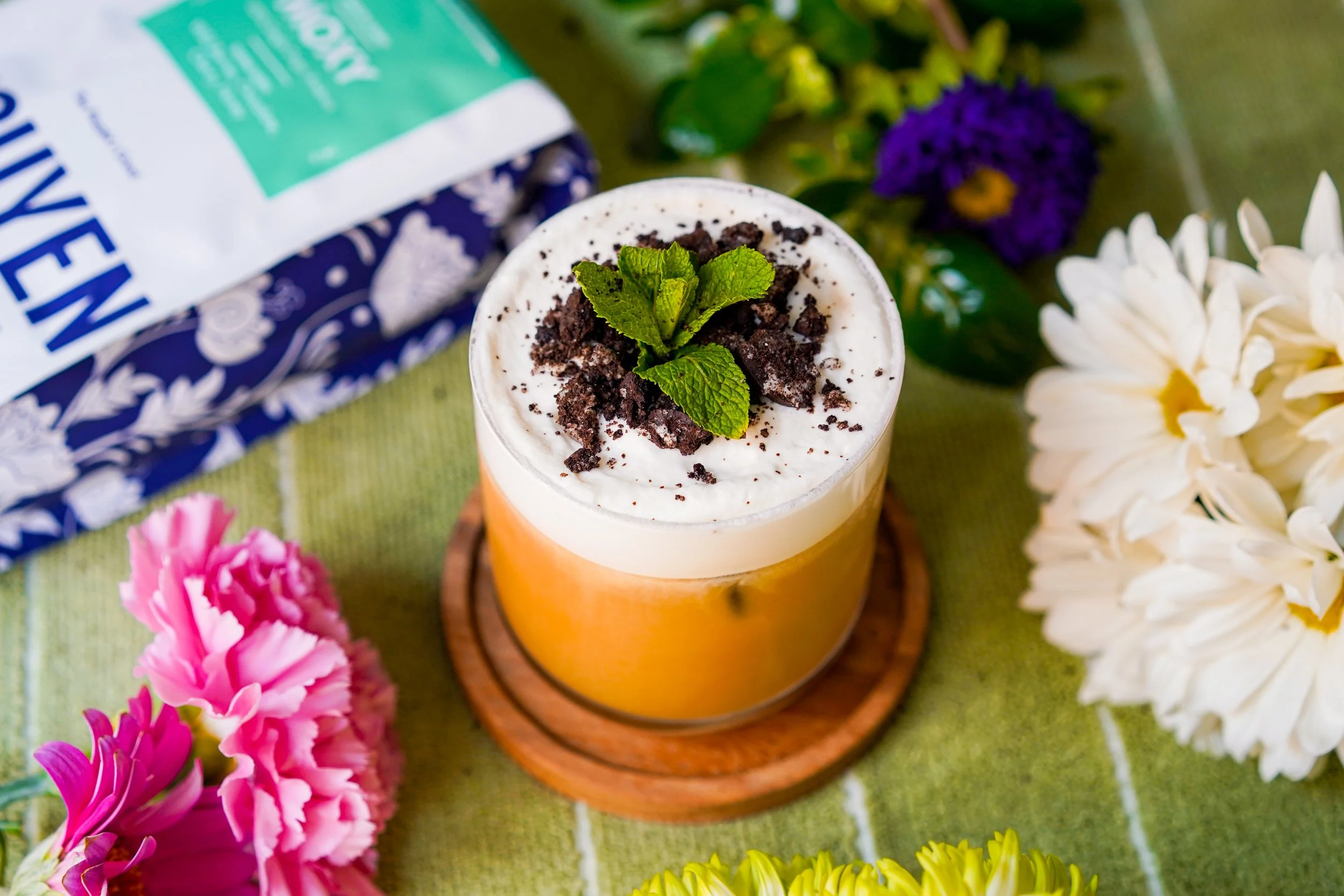 Iced coffee topped with whipped cream, crushed cookies, and mint on a coaster, surrounded by flowers and a coffee bag on a green surface.