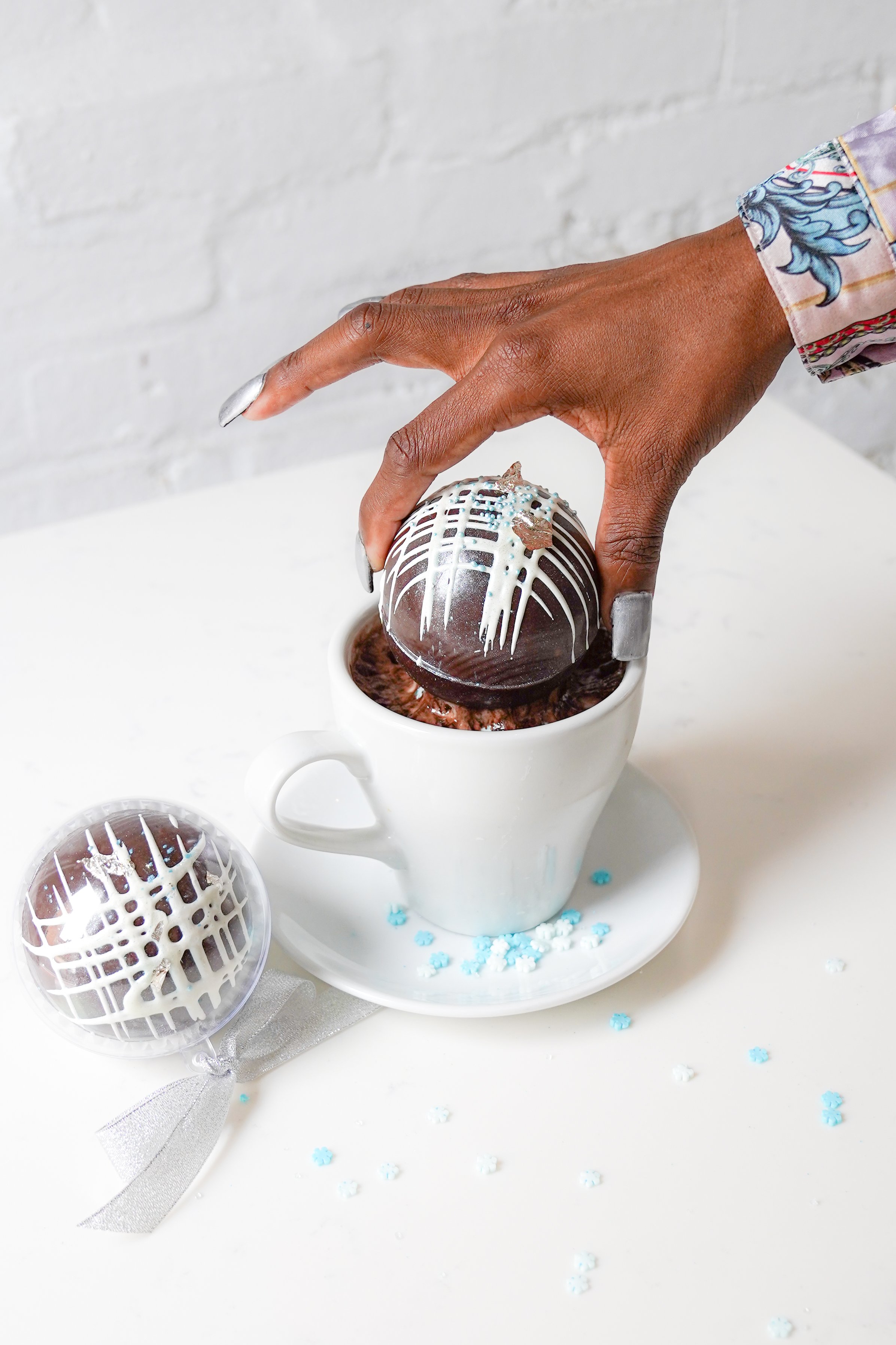 Person placing a decorative chocolate bomb in a cup of hot cocoa with another chocolate bomb beside it.