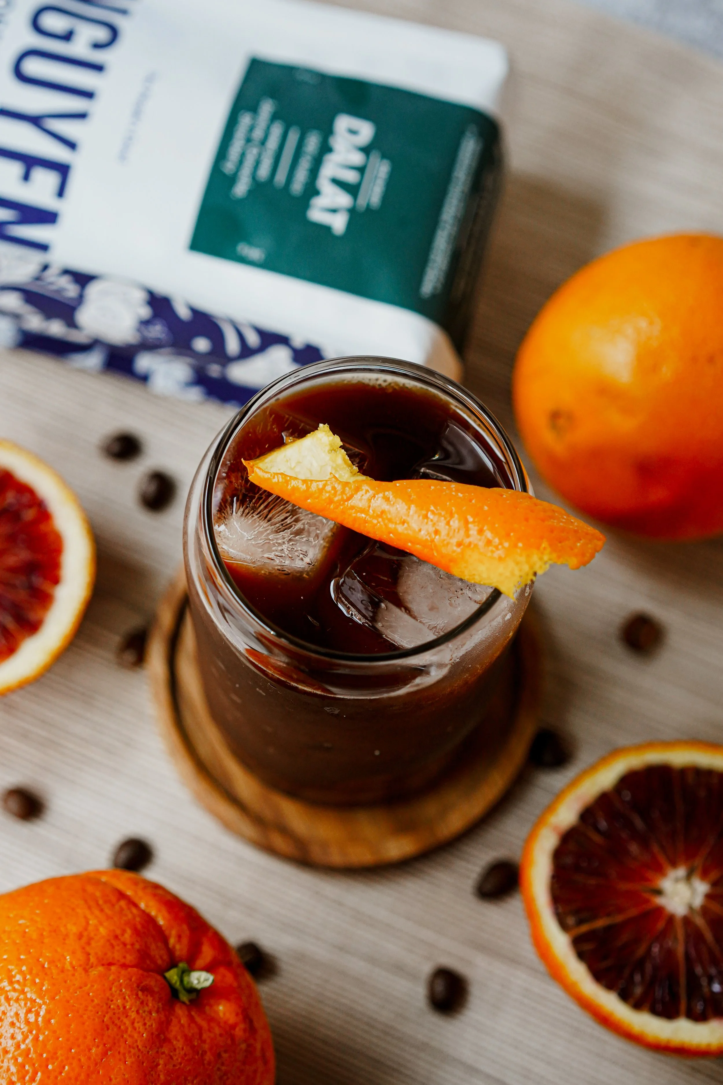 Iced coffee with orange slice garnish, whole and sliced oranges, coffee beans, and a Dalat coffee package on a table.