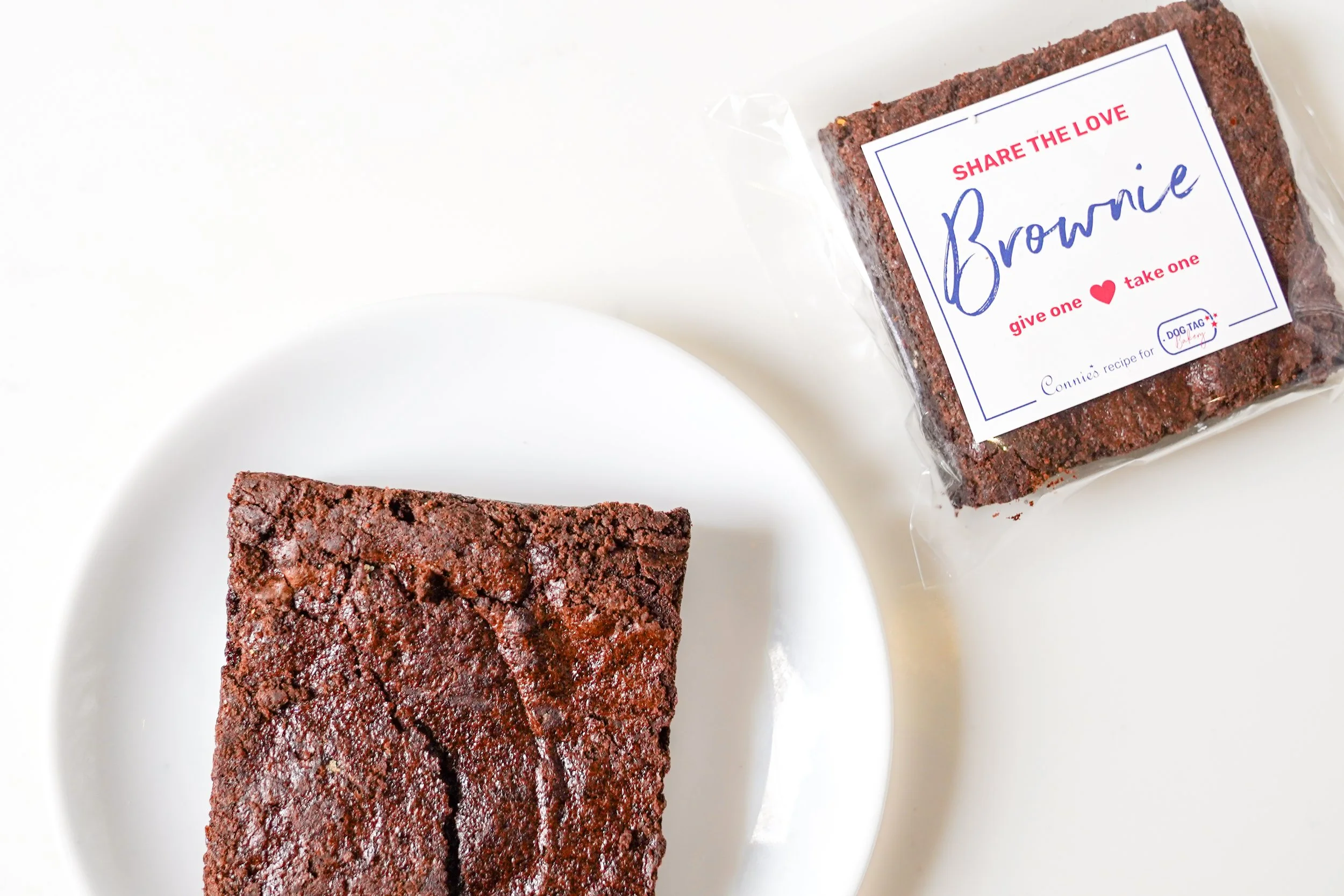 Chocolate brownie on a white plate with packaged brownie labeled 'Share the Love, Brownie, give one, take one.'