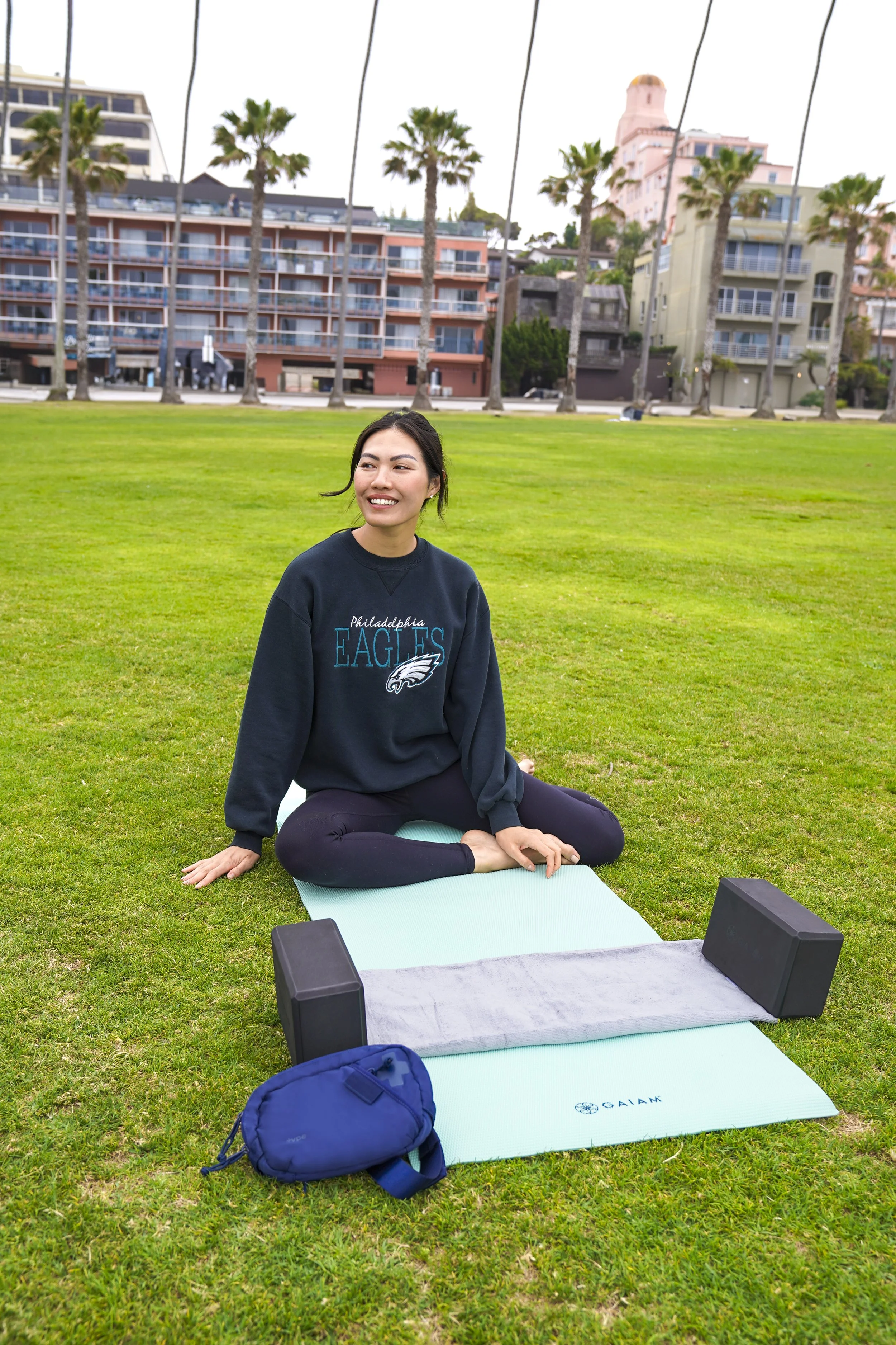 Woman sitting on yoga mat in park with yoga blocks and bag, wearing Philadelphia Eagles sweatshirt, buildings and palm trees in background.