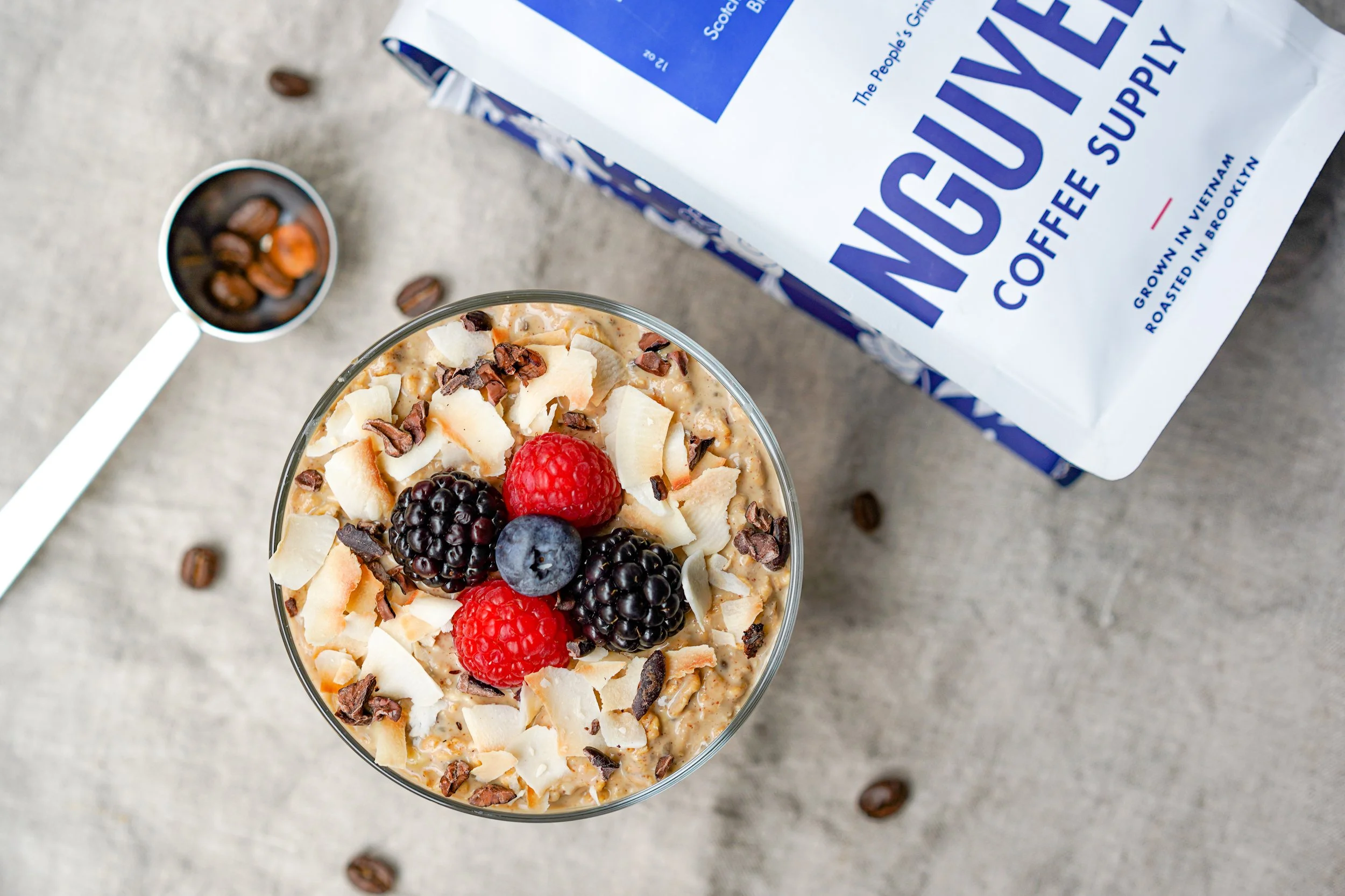 Bowl of oatmeal with berries, coconut flakes, and nuts, next to coffee package and spoon with beans.
