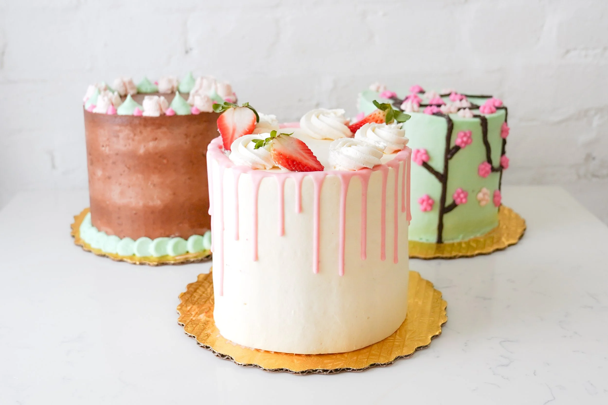 Three decorated cakes with different designs; one with pink and white icing drips and strawberries, another chocolate with mint green decorations, and the third with green icing and pink flowers.