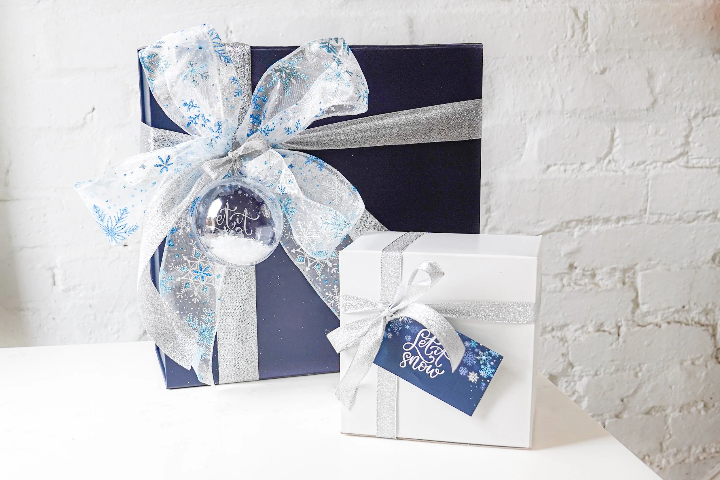 Two gift boxes with silver and blue snowflake-themed ribbons and tags on a white surface.