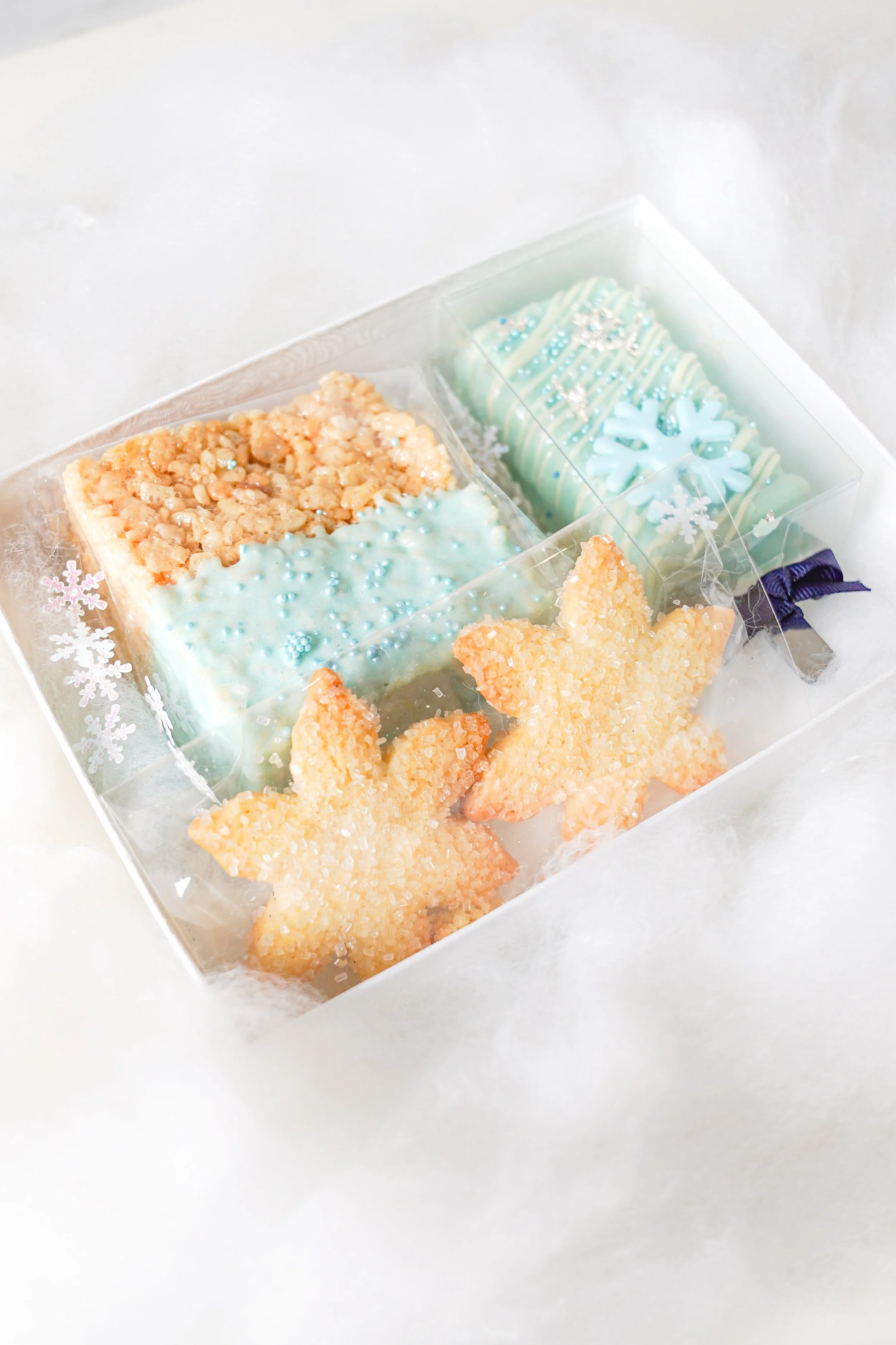 Winter-themed treats in a box, including snowflake-shaped cookies and rice crispy treats with light blue icing and sprinkles.