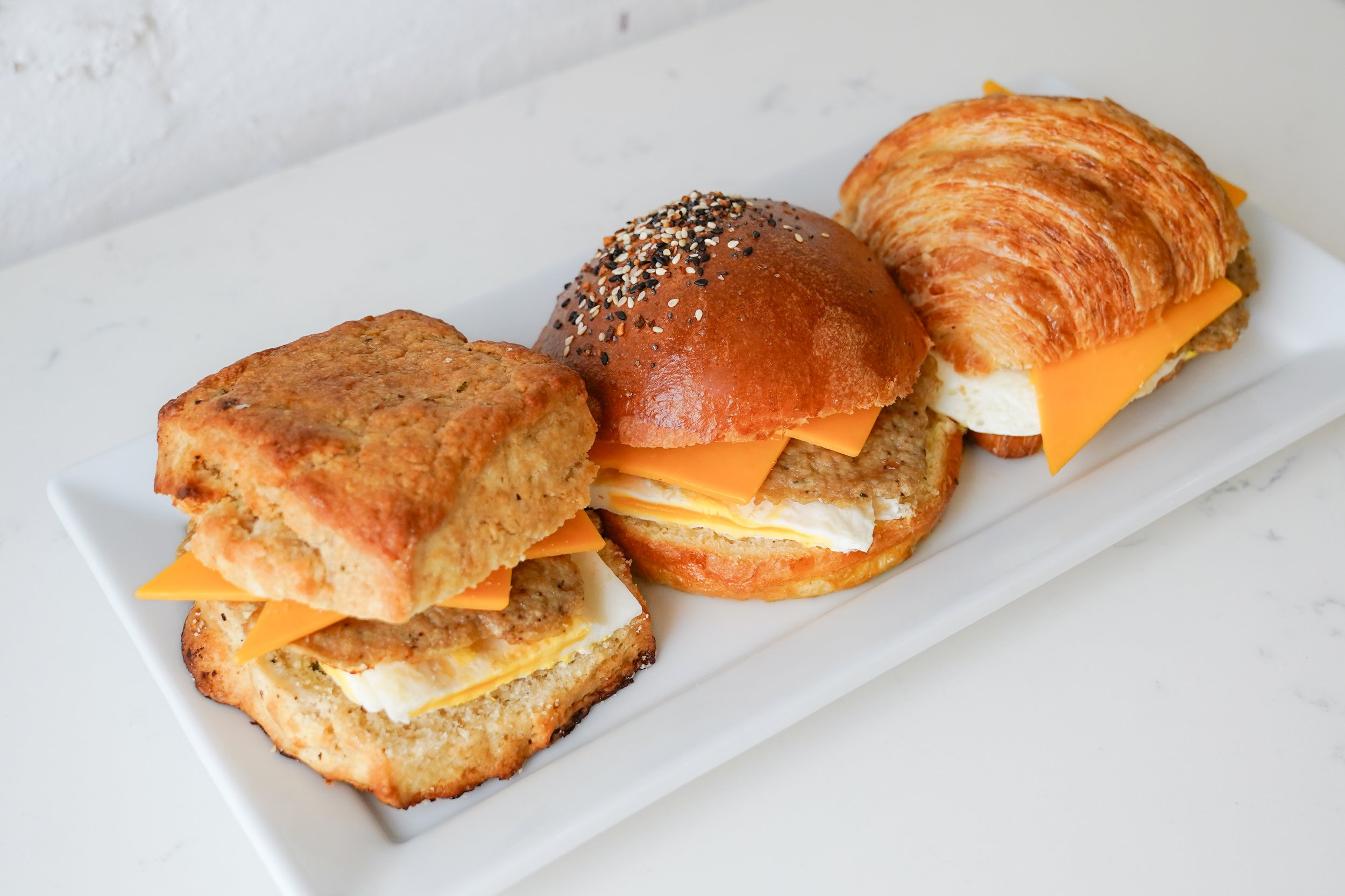Three breakfast sandwiches on a white plate with different breads: biscuit, sesame bun, and croissant, each filled with cheese, sausage, and egg.