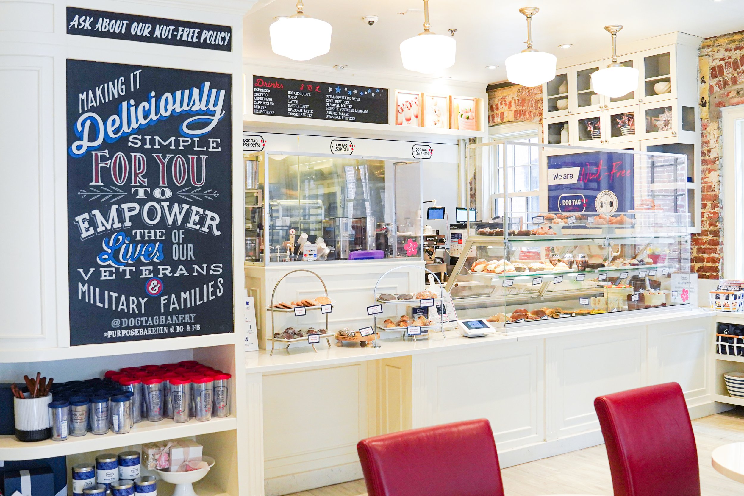 Interior of a bakery with a sign promoting support for veterans and military families, display cases with pastries, a coffee menu, and red chairs.
