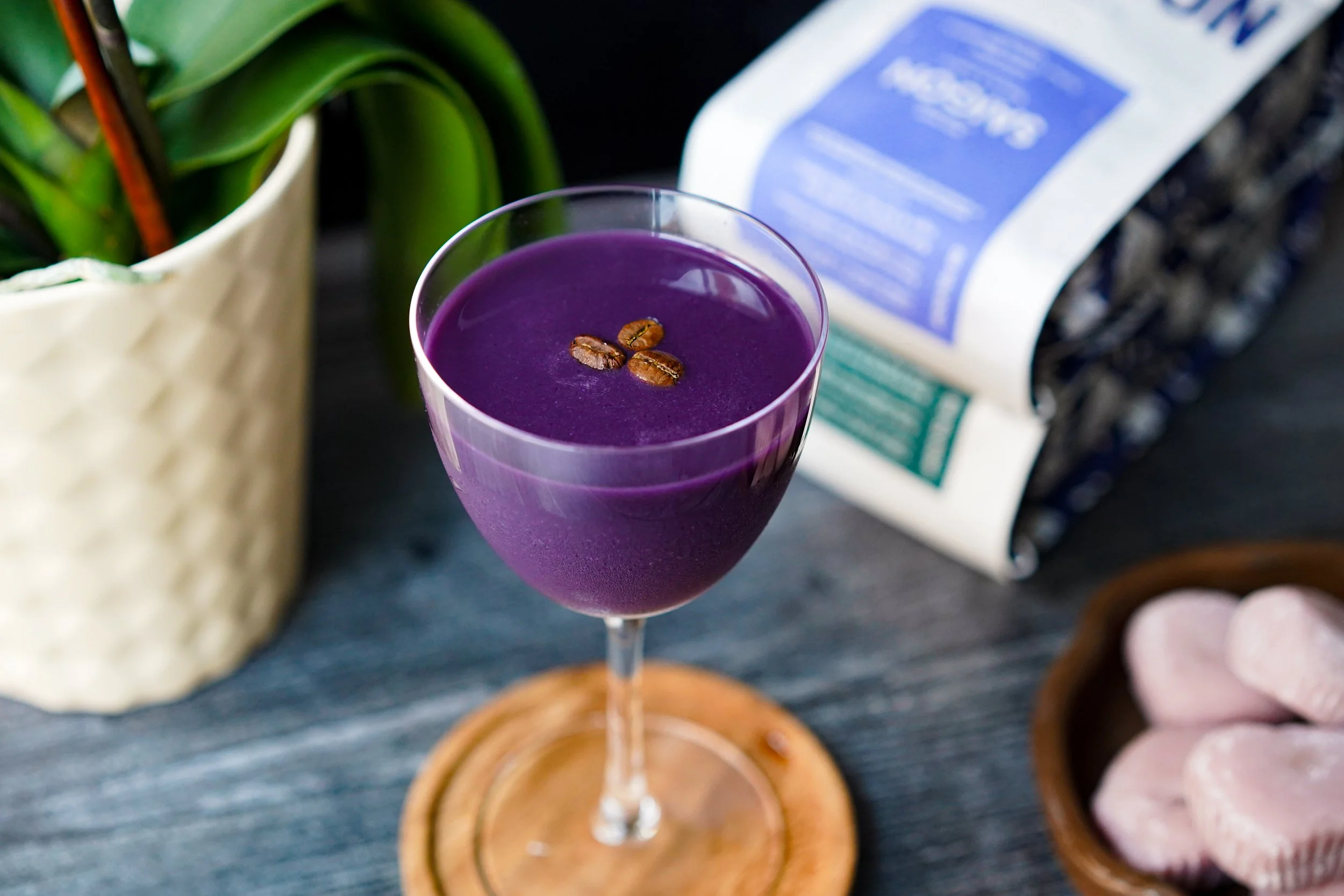 Purple cocktail with coffee beans garnish on wooden coaster, next to a plant and pastries.