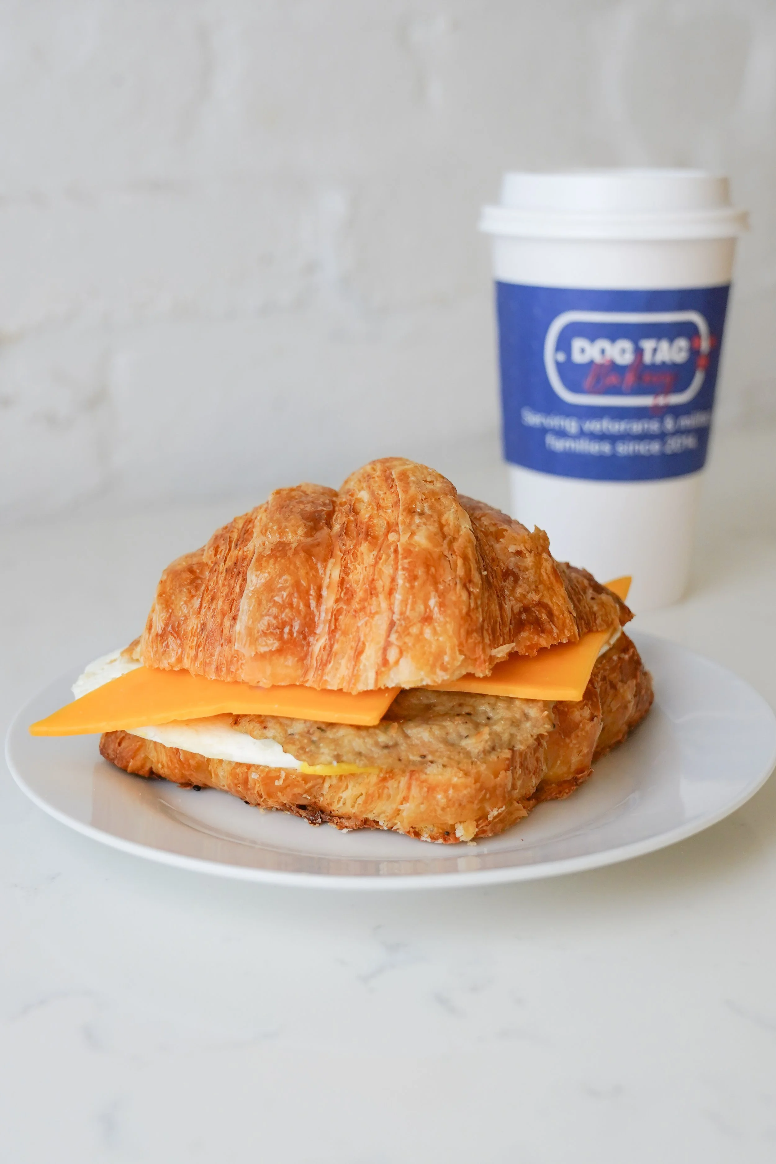 Croissant sandwich with cheese and sausage on a plate, coffee cup in background.