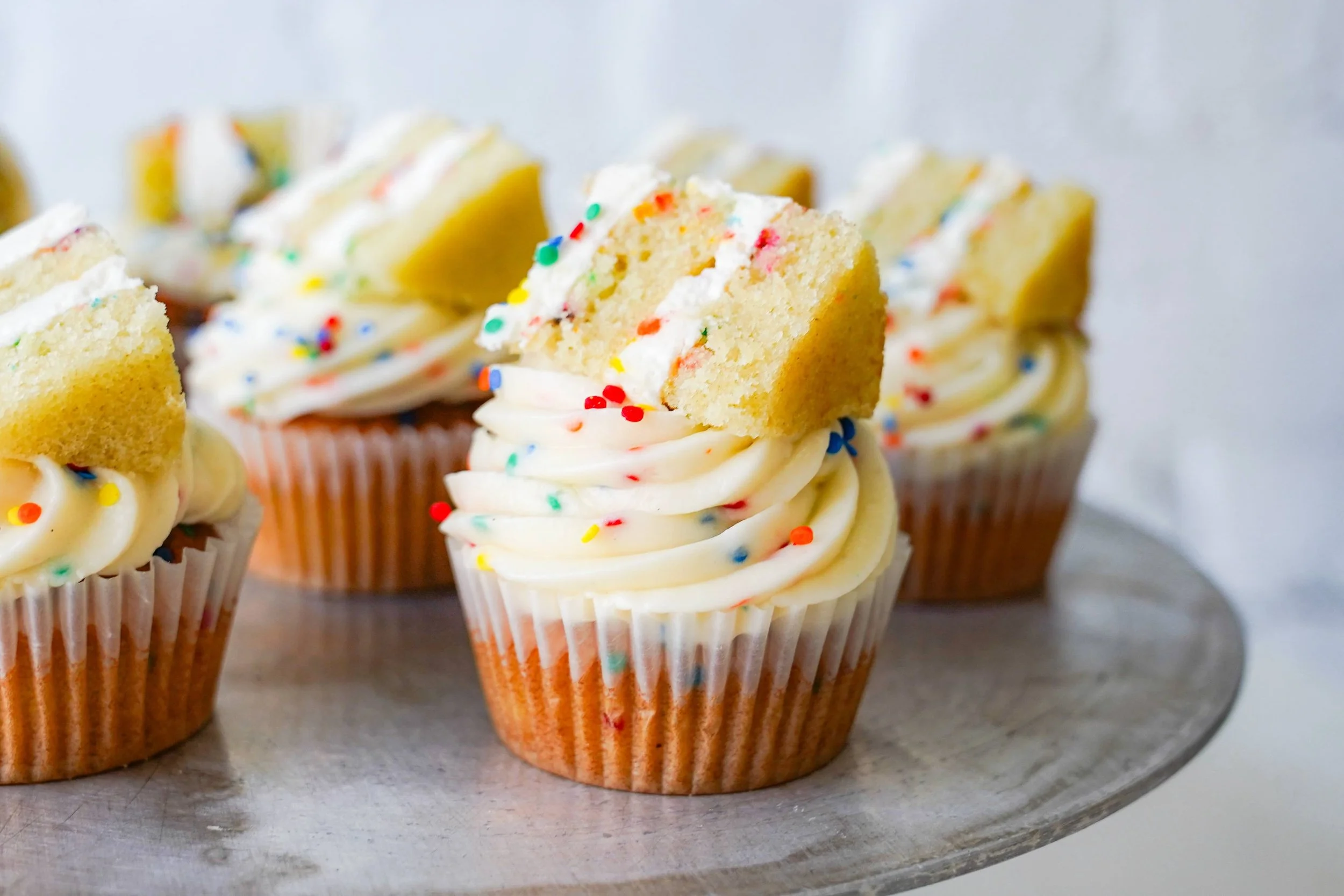 Cupcakes with white frosting, rainbow sprinkles, and cake slices on top, displayed on a tray.