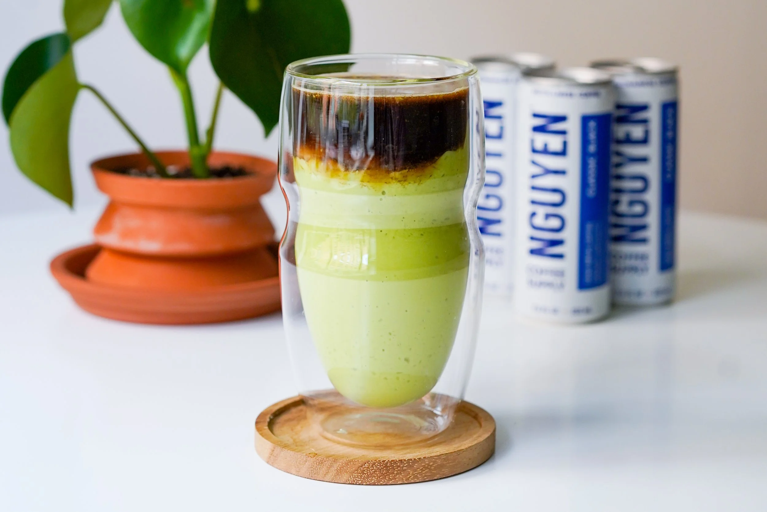 Layered matcha and coffee drink in glass cup on wooden coaster, with potted plant and Nguyen coffee cans in background.