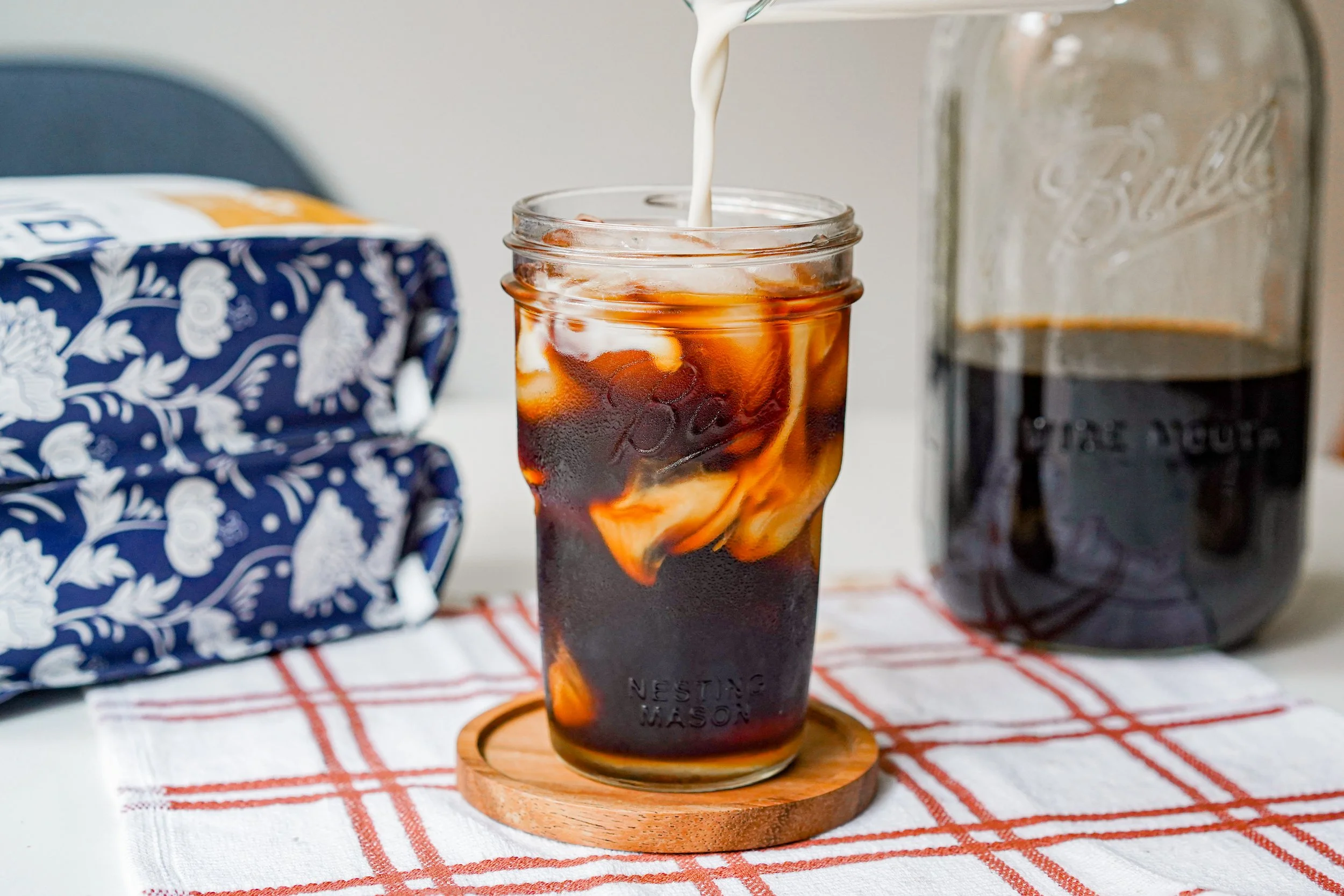 Iced coffee in a mason jar with milk being poured, placed on a wooden coaster on a plaid cloth, next to a jar of coffee and a blue patterned item.