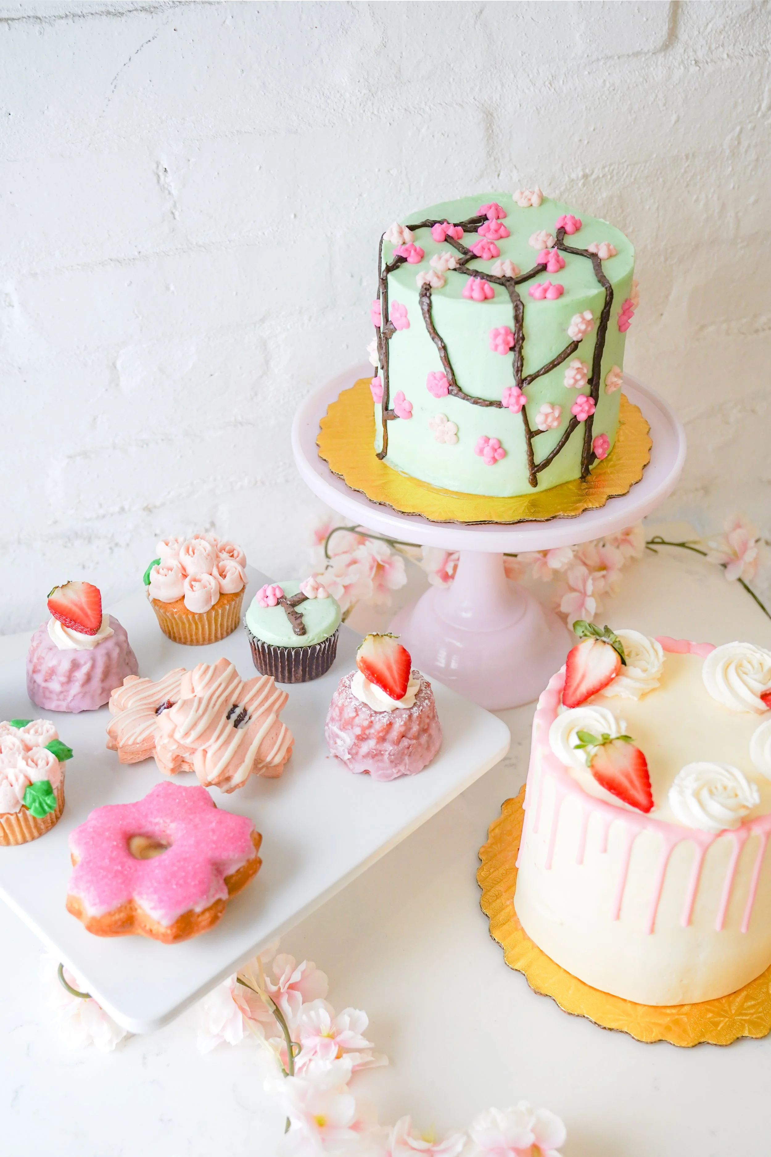 Assorted pastel cakes and cupcakes with floral decorations and strawberries, displayed on a tabletop against a white background.